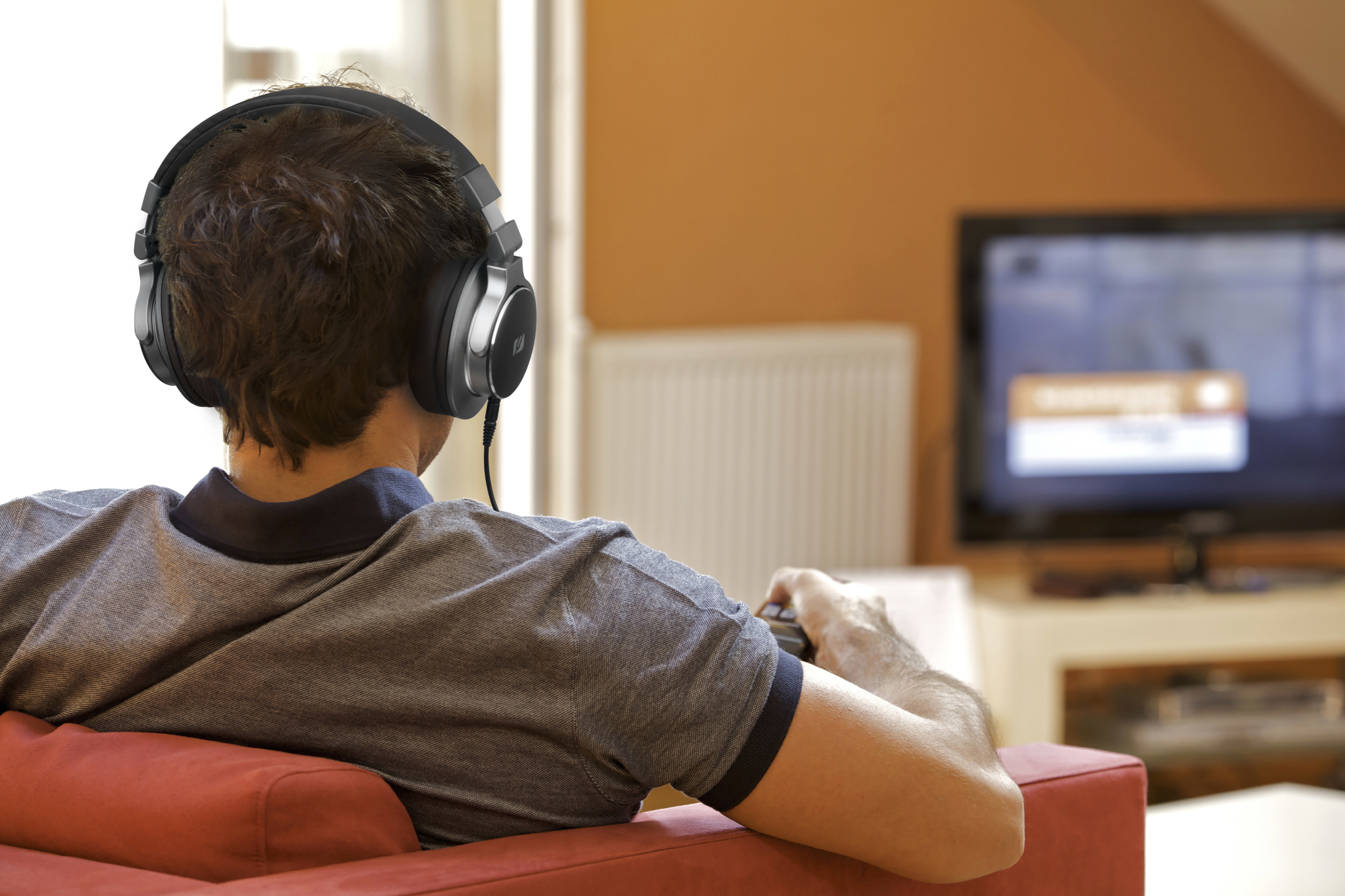 Uomo con cuffie, guarda la TV. Divano rosso, muro arancione. Tiene un controller. Focus sulla nuca.