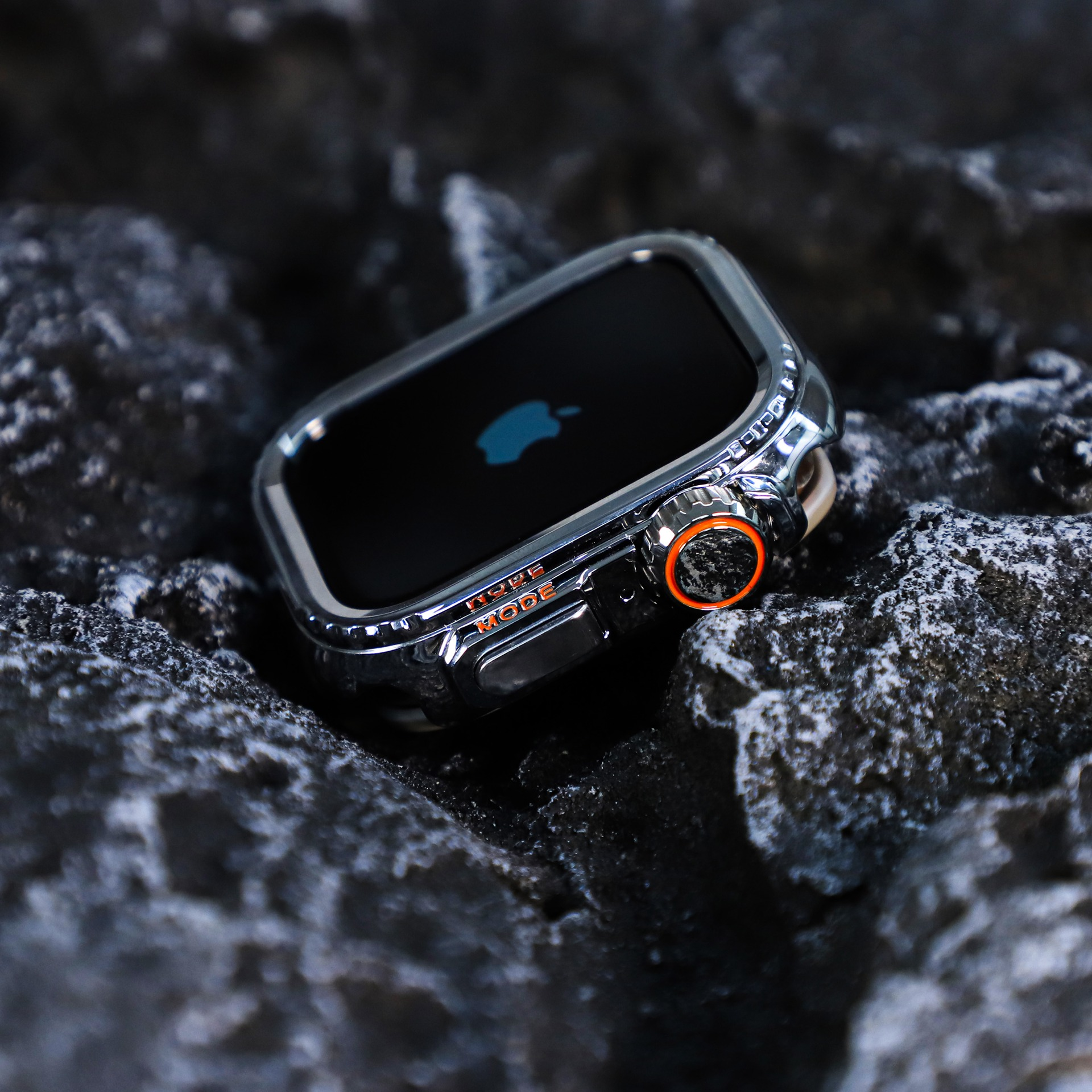 Uno smartwatch argentato con un pulsante arancione, appoggiato su rocce scure e strutturate. Il quadrante mostra il logo Apple.