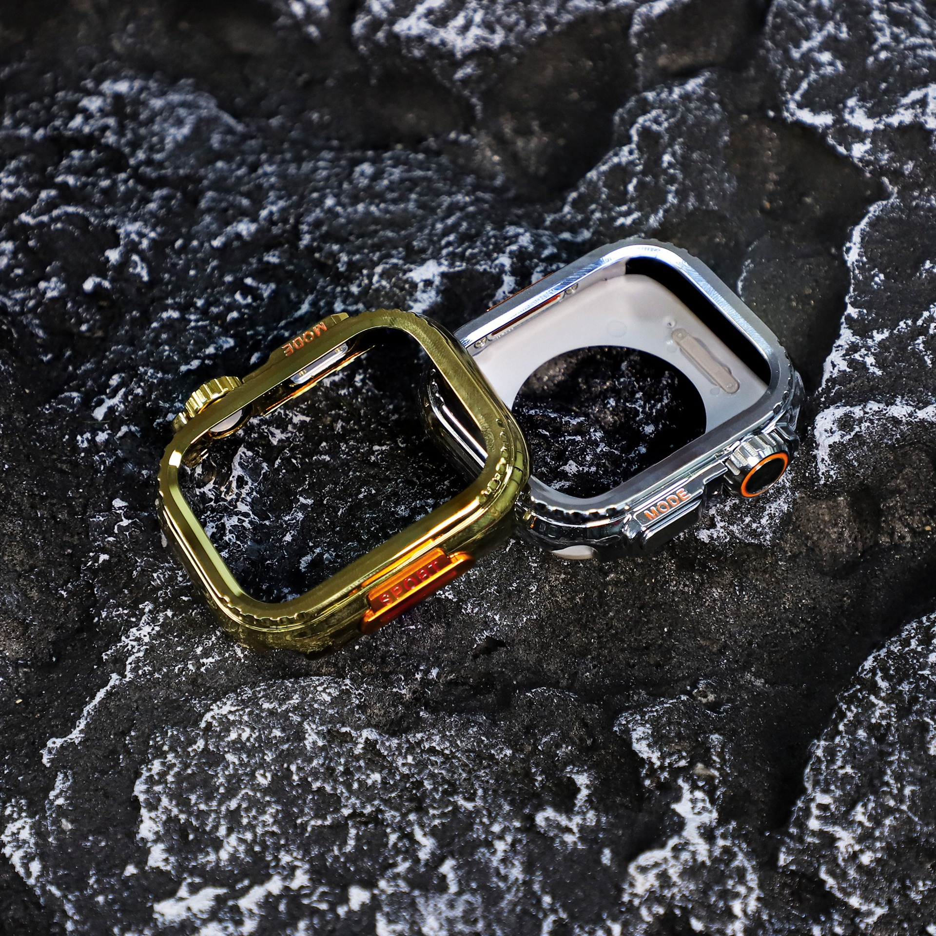 Due custodie per orologi, oro e argento, su una superficie rocciosa nera. La custodia dorata ha dettagli arancioni. La custodia argentata è accanto.