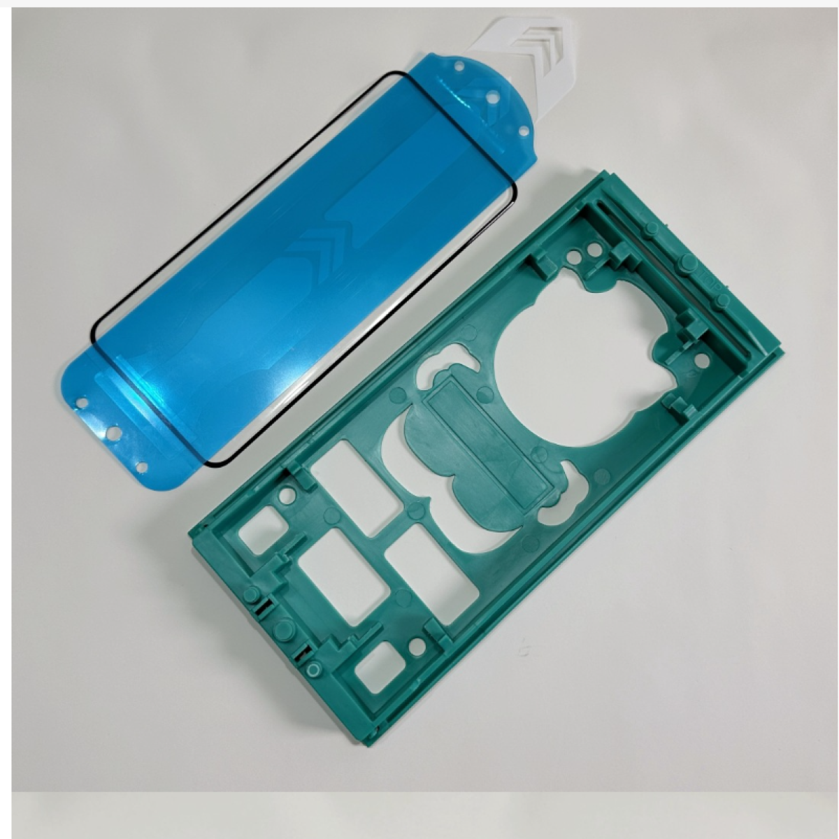 Dispositivo smontato con protezione schermo blu, cornice verde e pezzo di plastica bianco su superficie bianca.