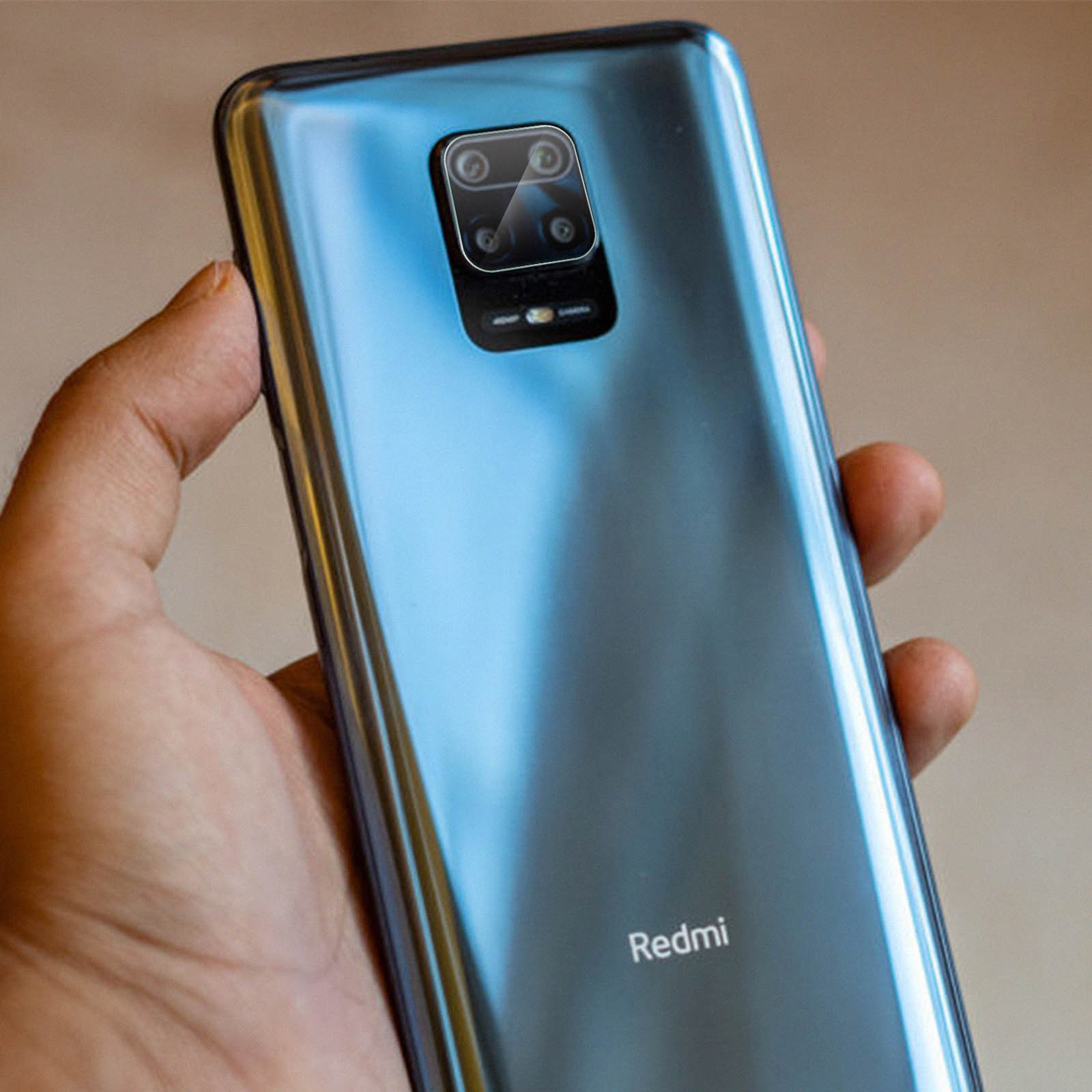 Une main tient un téléphone Redmi bleu. La caméra du téléphone est visible. Le fond est flou et clair.