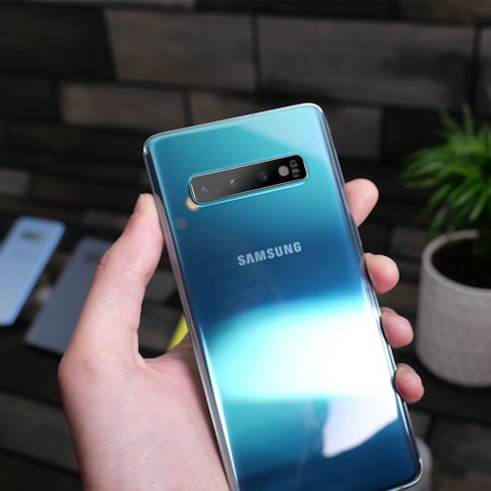 Una mano tiene un telefono Samsung verde acqua. Lo sfondo ha un muro di mattoni, altri telefoni e una pianta.