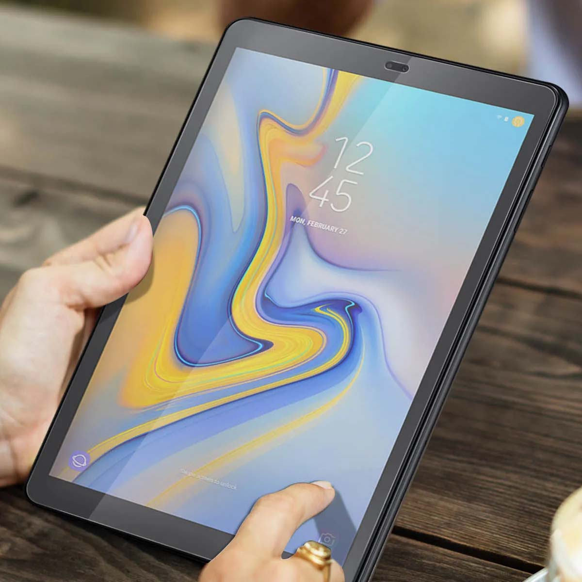 Een persoon houdt een zwarte tablet vast met 12:45, maandag 27 februari. Het scherm heeft een geel en blauw abstract ontwerp.