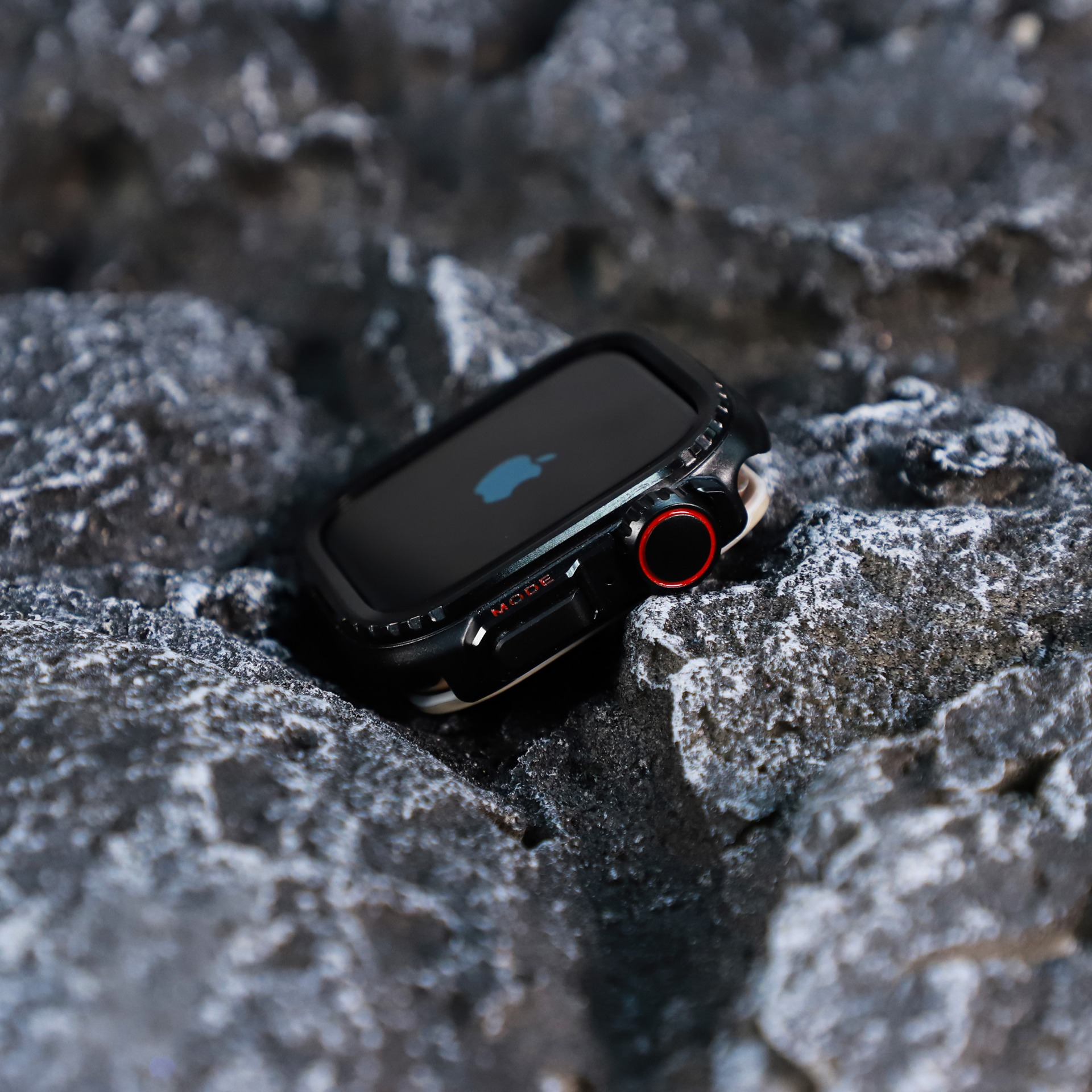 Apple Watch nera su una roccia grigia strutturata. L'orologio ha una lunetta nera e un quadrante rosso. Logo Apple visibile.