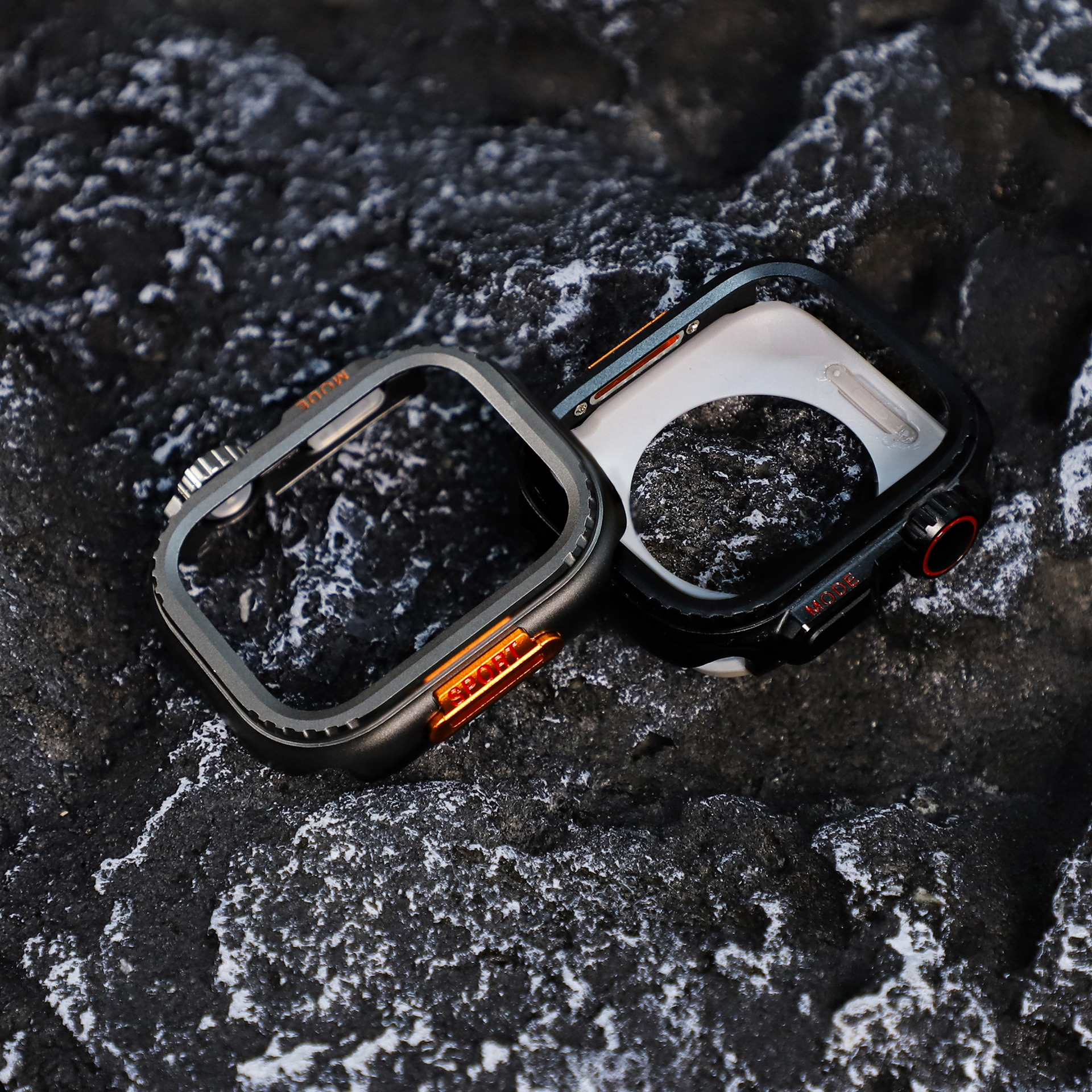 Due custodie per orologi su una superficie nera strutturata. Una grigia con dettagli arancioni, l'altra nera con rosso.