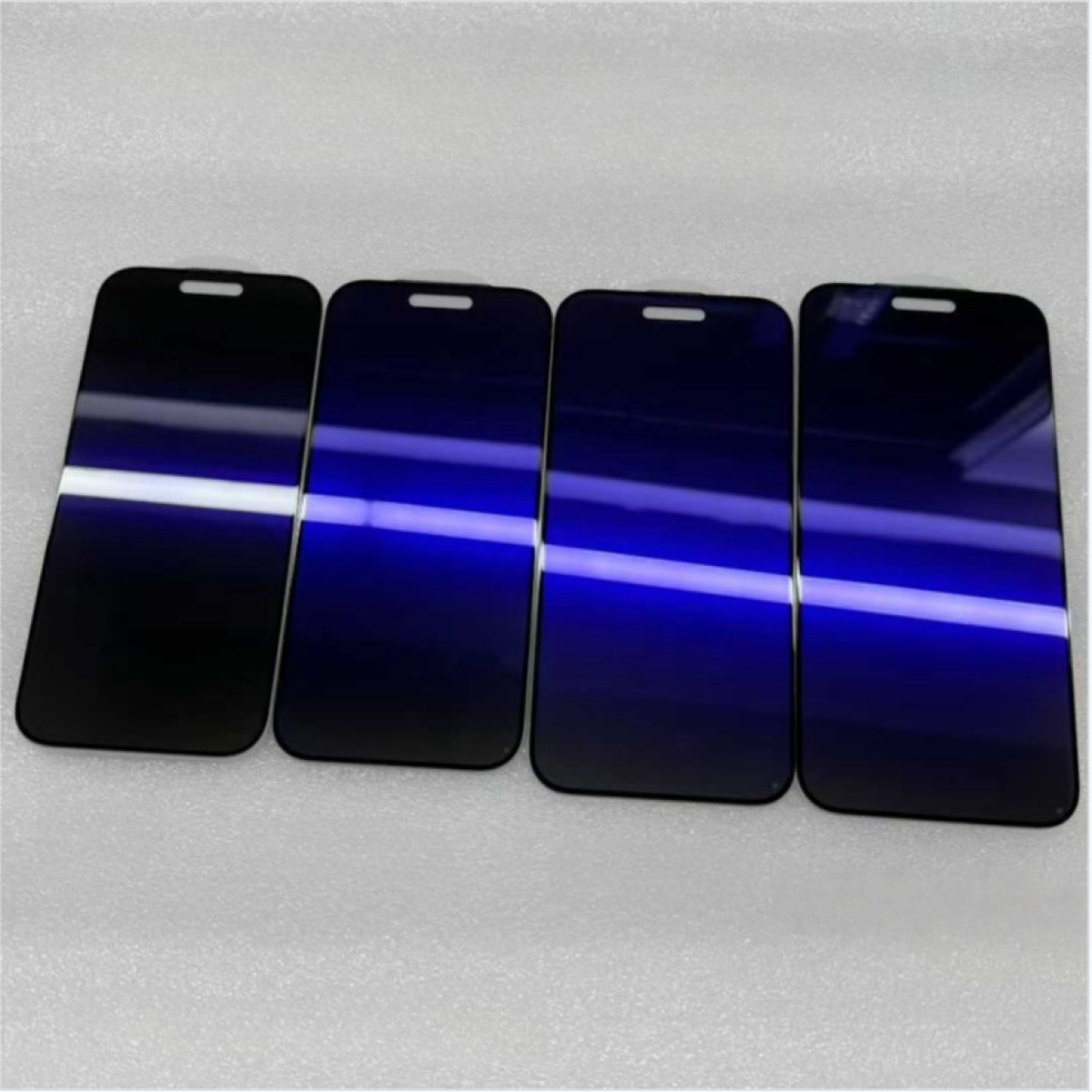 Quattro protezioni schermo blu scuro per telefoni disposte fianco a fianco su una superficie bianca, riflettendo la luce.
