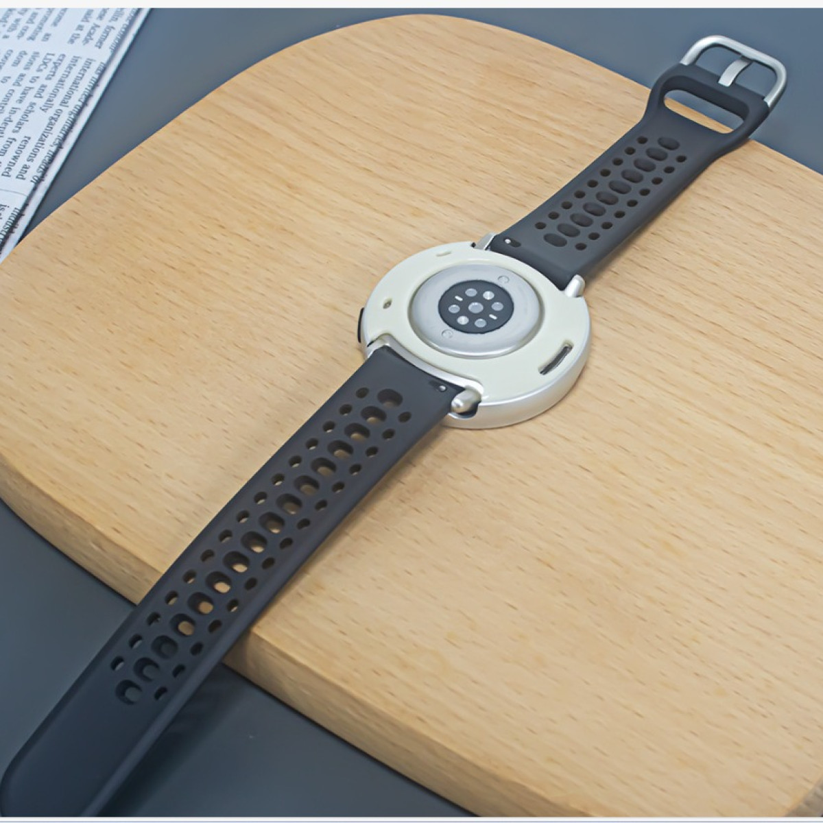 Uno smartwatch con cinturino nero, posto su un tagliere di legno. L'orologio ha cassa argentata e sensore sul retro.