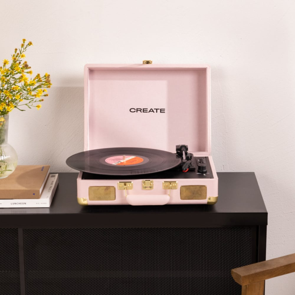 Tocadiscos rosa sobre una mesa negra con libros y flores en un jarrón. La palabra 'CREATE' está en el reproductor.