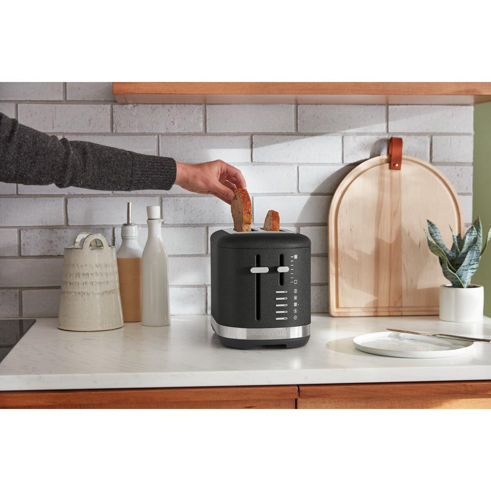 Eine Hand legt Toast in einen schwarzen Toaster. Küchenzeile mit Flaschen, Schneidebrett und einer Pflanze im Hintergrund.