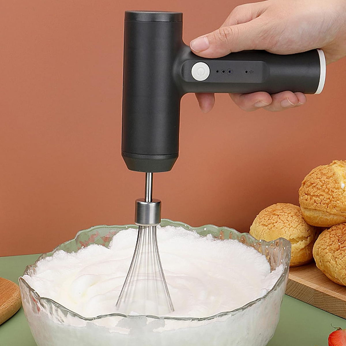 Eine Hand hält einen schwarzen Mixer über einer Schüssel mit Schlagsahne. Gebäck liegt rechts.