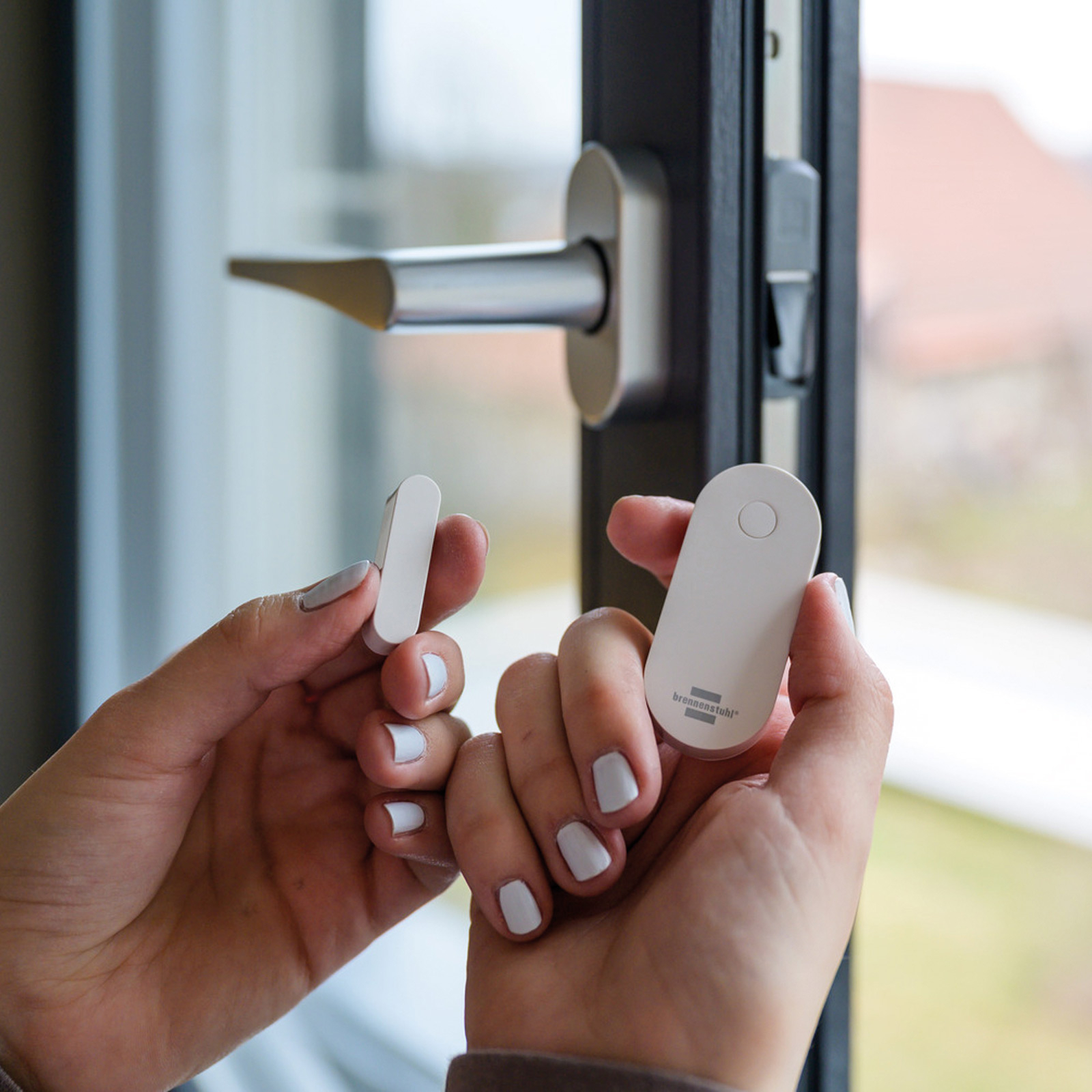 Hände halten weiße Sensoren und einen Türgriff. In jeder Hand wird ein Sensor gehalten, in der Nähe eines Fensterrahmens.