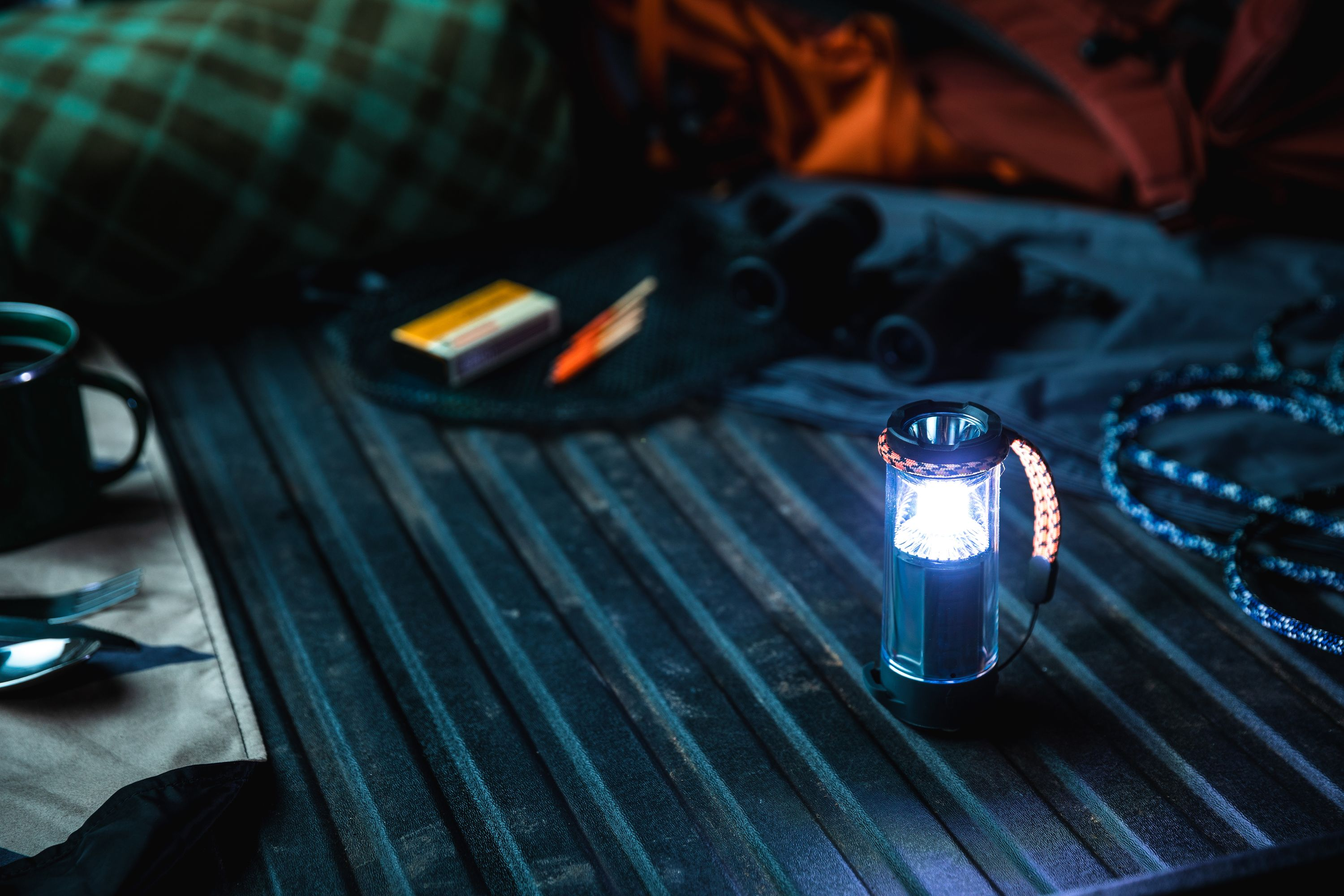 Una linterna encendida sobre una superficie oscura, con equipo de acampada visible en el fondo, incluyendo taza y utensilios.