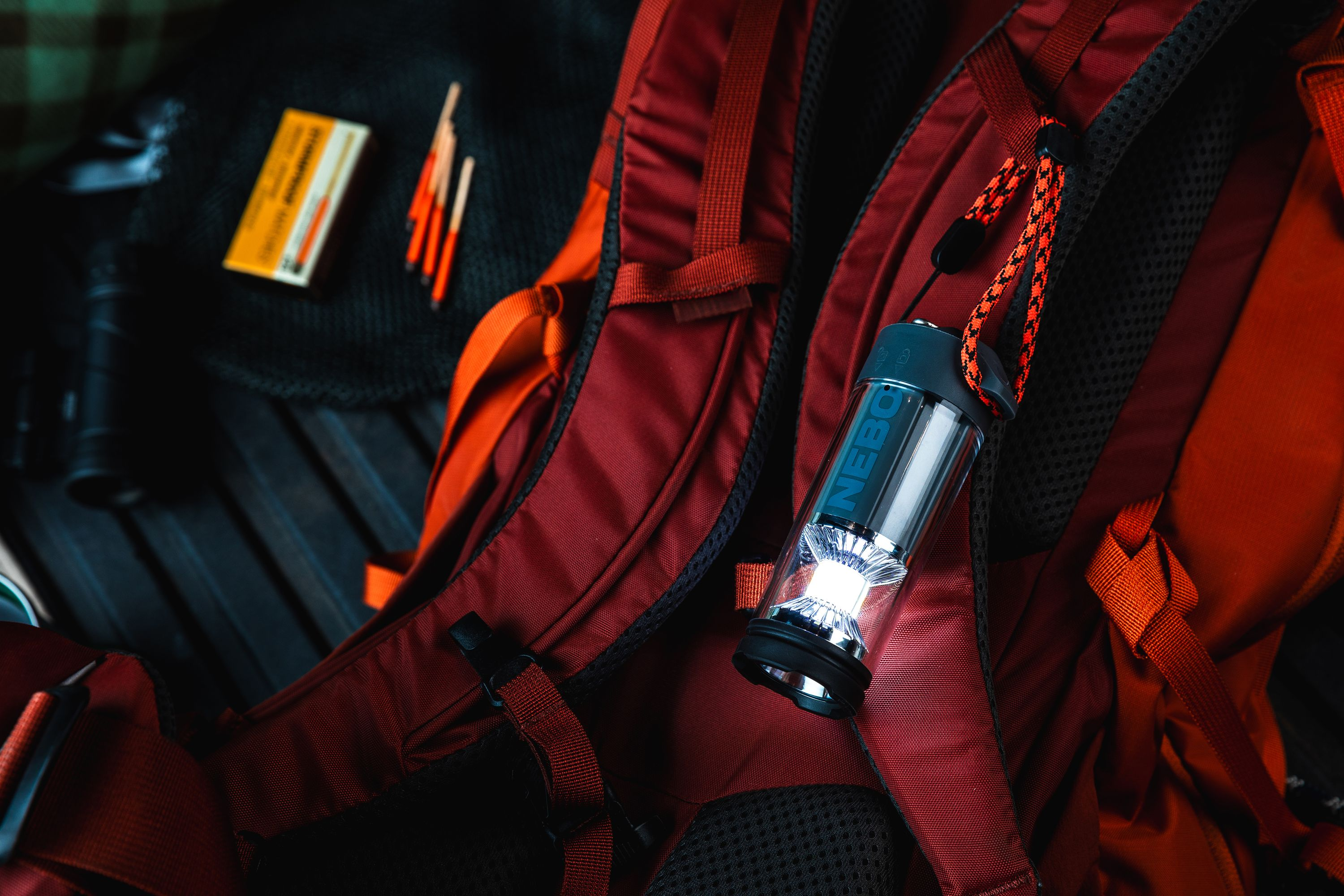 Una mochila roja con una linterna. Fósforos y una bolsa negra encima. Fondo de madera oscura.