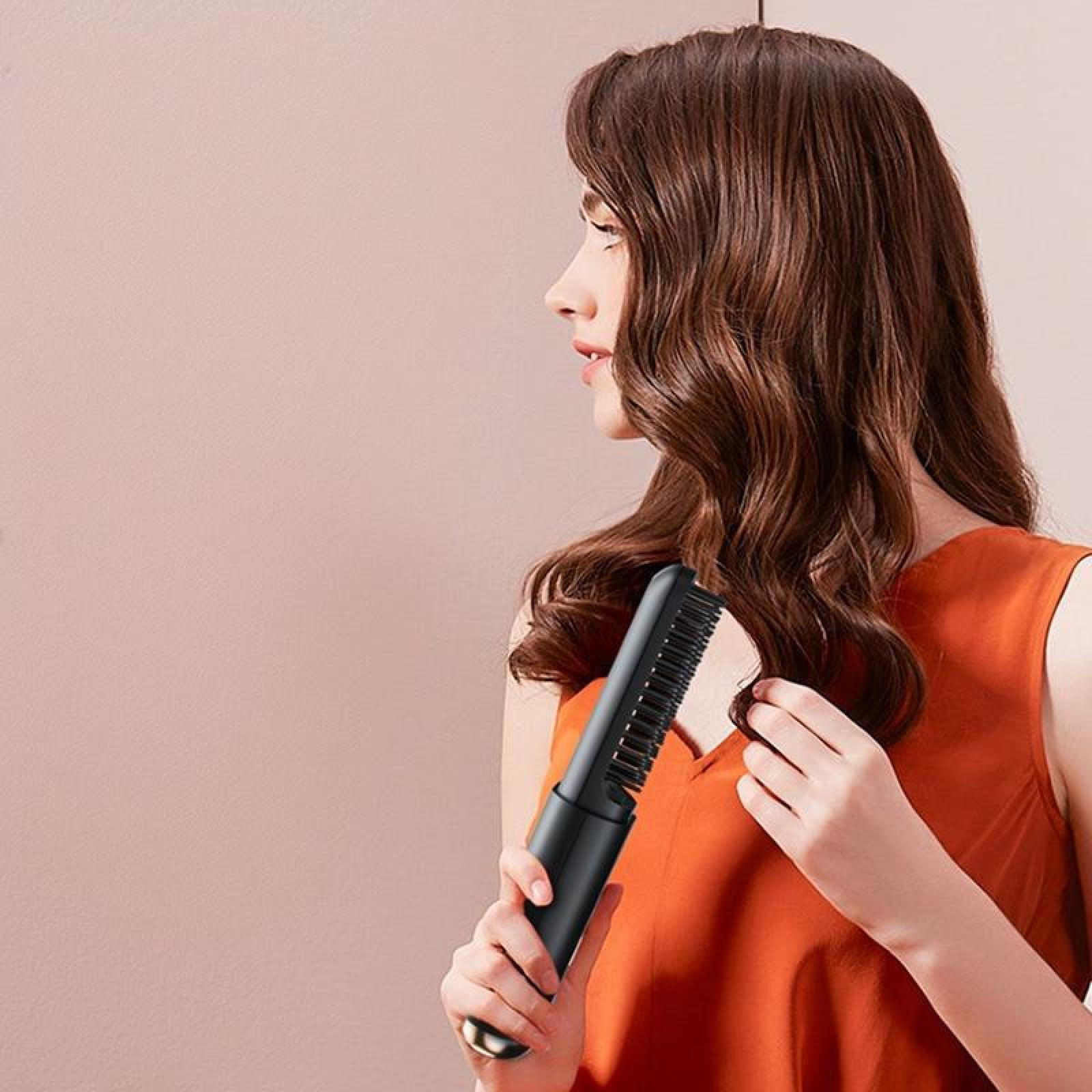 Eine Frau benutzt einen schwarzen Haarglätter. Sie trägt ein orangefarbenes Oberteil, rosa Hintergrund. Braune Haare.