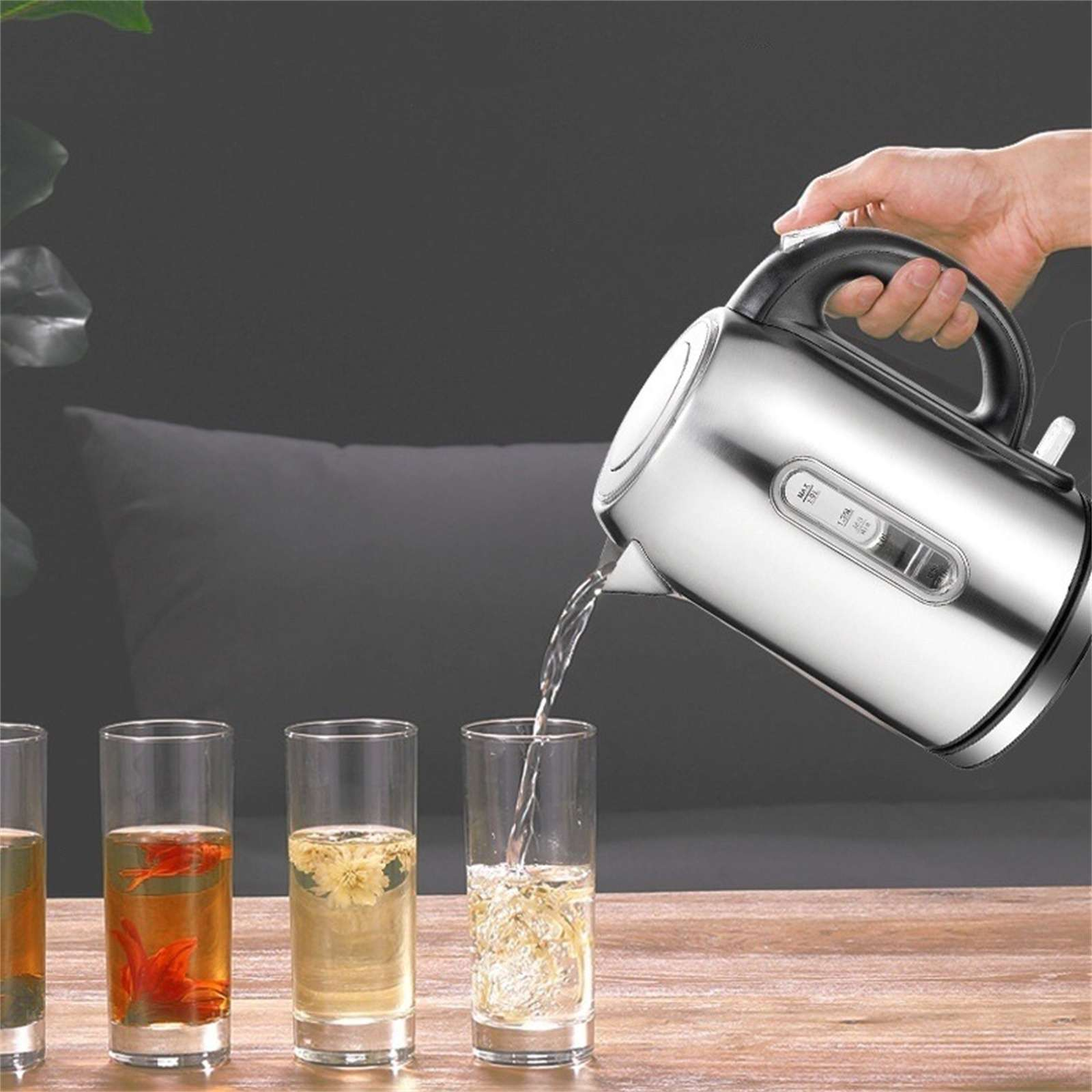 Eine Hand gießt Wasser aus einem silbernen Wasserkocher in ein Glas. Drei Gläser mit Tee stehen auf einem Holztisch.