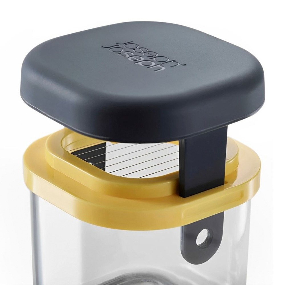 Primo piano di un utensile da cucina. Ha un coperchio grigio scuro con il marchio. Sotto, un telaio giallo con lame e un contenitore trasparente.