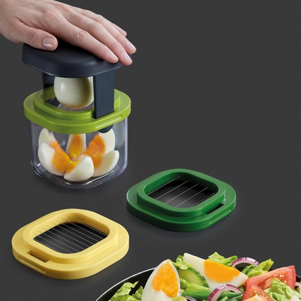 Una mano preme su un tagliauova. Uova a fette sono in un contenitore. Sono presenti anche due affettatrici e una ciotola di insalata.