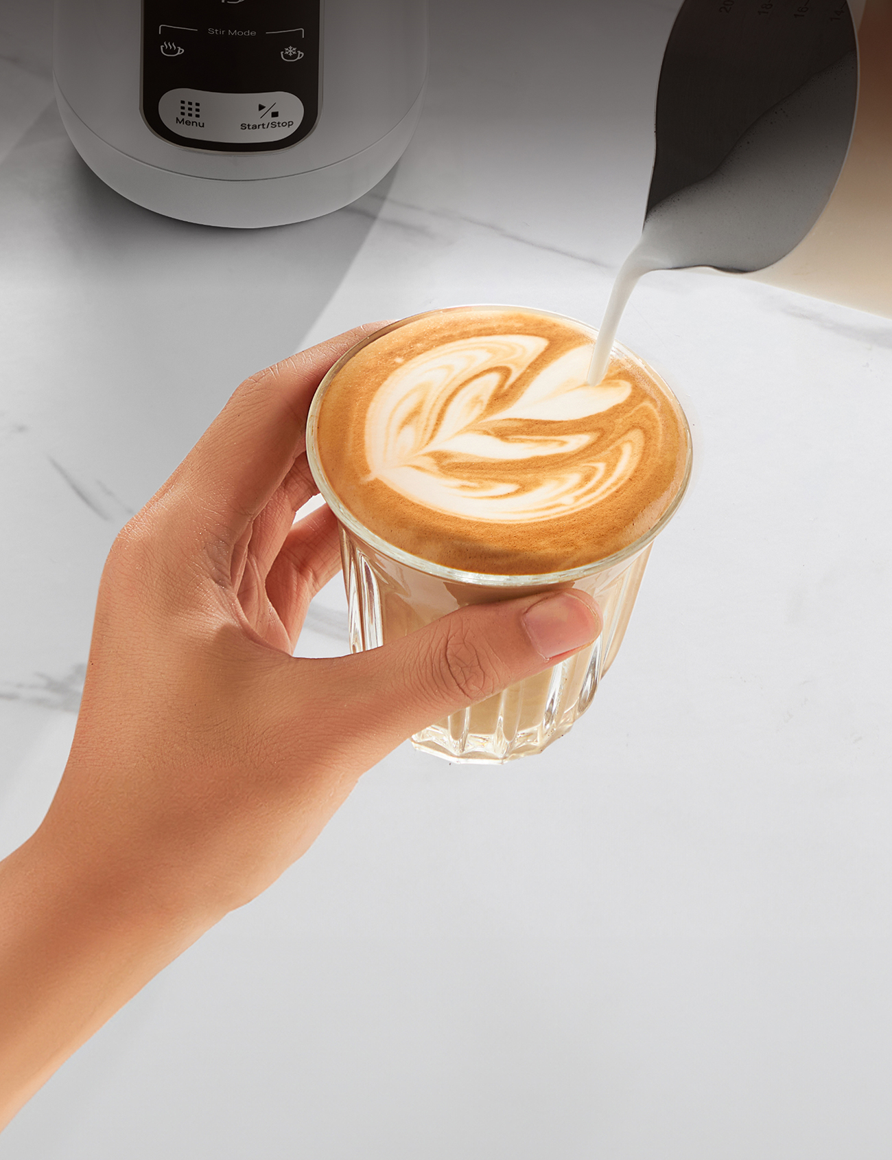 Hand gießt Milch in einen Latte mit Blattmuster. Glasbecher wird von Hand gehalten. Weiße Arbeitsplatte und Kaffeemaschine.