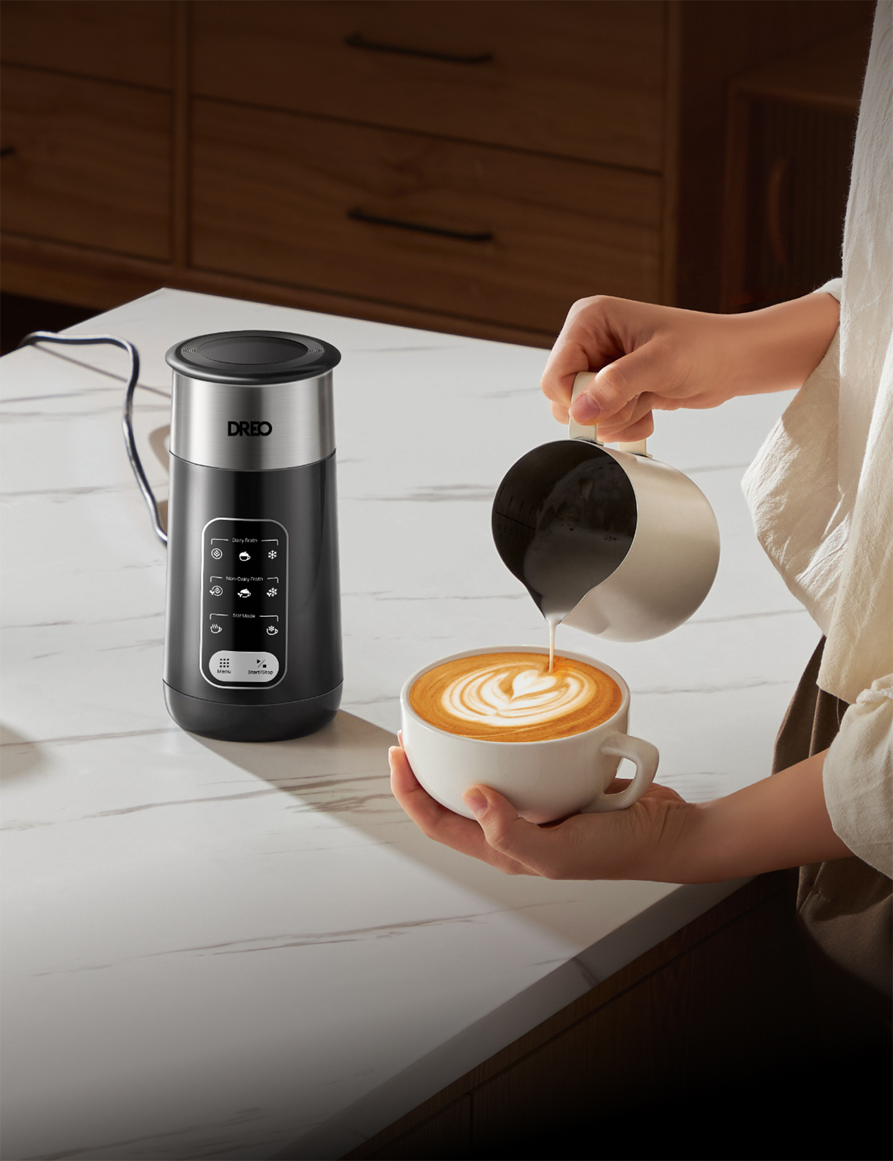 Eine Person gießt Milch aus einem Krug in eine Kaffeetasse mit Latte Art, links steht ein schwarzer und silberner Aufschäumer.
