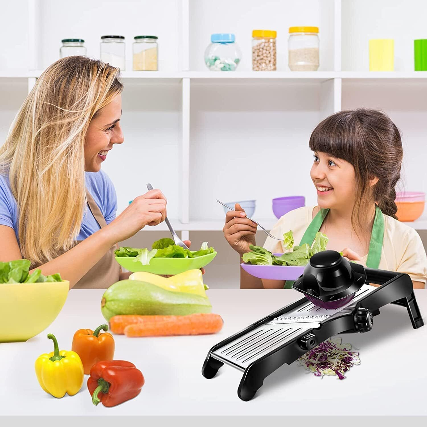 Eine Frau und ein Mädchen bereiten in einer Küche Salat zu, wobei sie einen Schneider verwenden. Gemüse wie Paprika, Karotten und Zucchini liegen auf dem Tisch.
