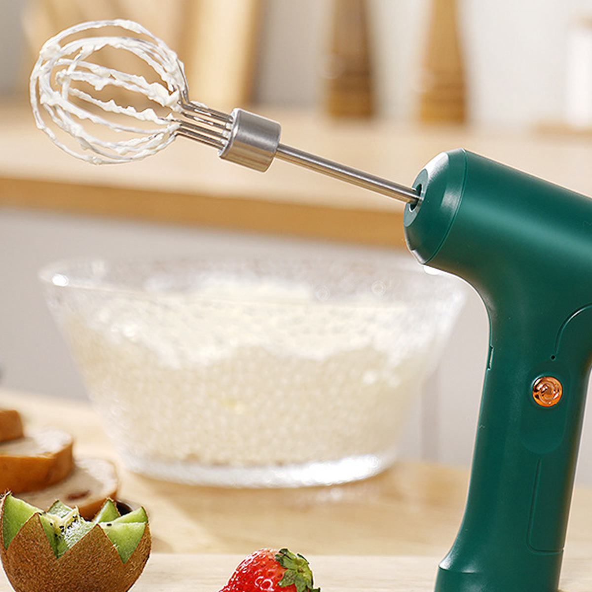 Ein grüner elektrischer Mixer mit Schneebesen verrührt Sahne in einer klaren Schüssel. Kiwi und Erdbeere auf dem Tisch.