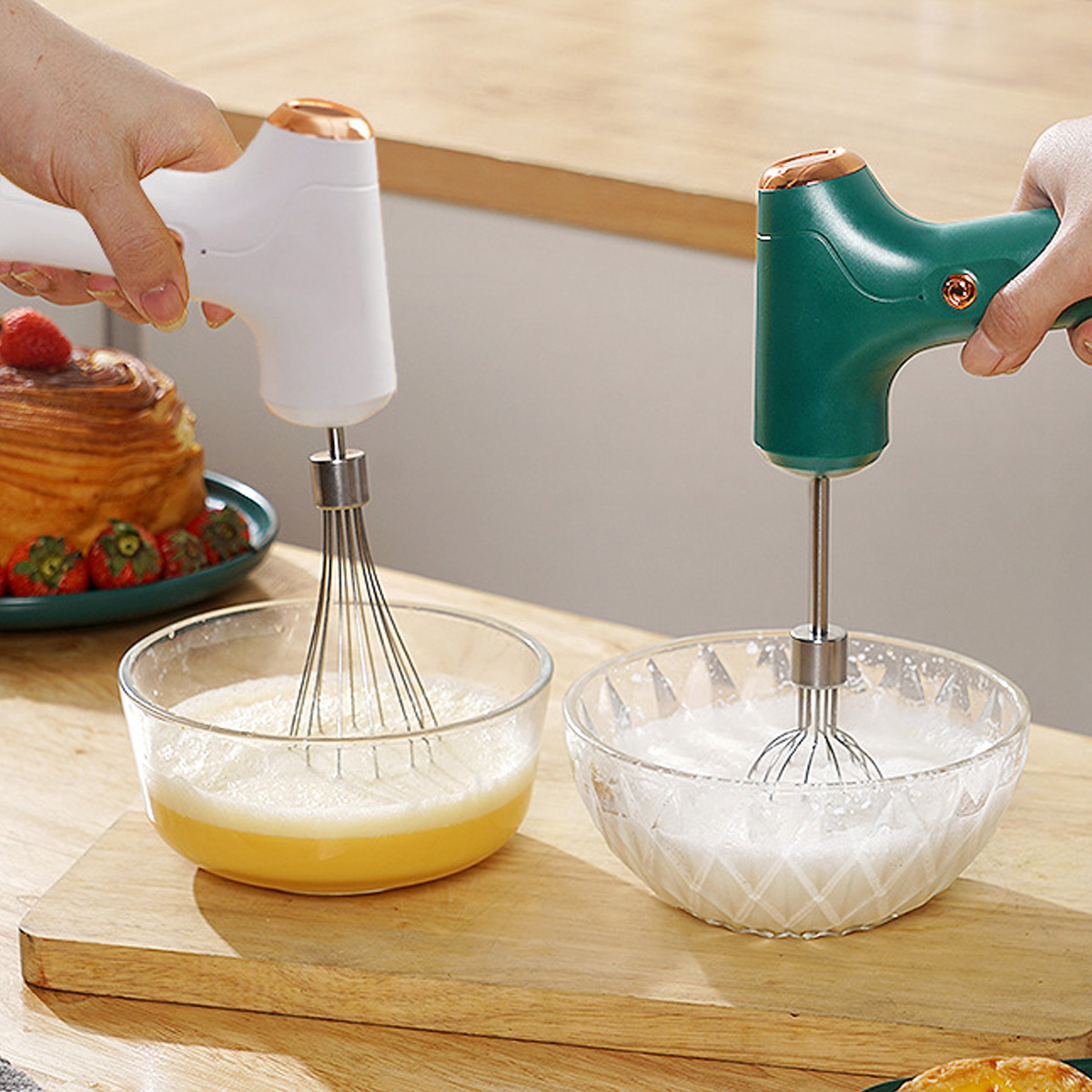 Zwei Hände halten Mixer über Schüsseln mit Zutaten. Eine Schüssel hat gelbe Flüssigkeit, die andere weiß. Kuchen und Erdbeeren sind im Hintergrund.
