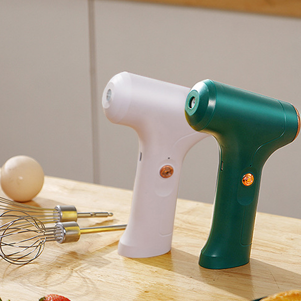 Zwei Handmixer, weiß und grün, stehen auf einer Holzoberfläche mit einem Schneebesen und einem Ei. Weicher Hintergrund.