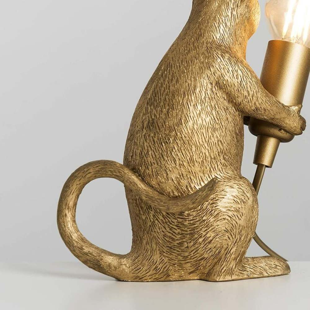 Eine goldene Affenlampe. Der Affe hält eine Glühbirne. Der Schwanz ist eingerollt. Der Hintergrund ist weiß.
