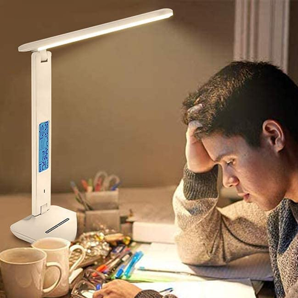 Ein Mann lernt unter einer weißen Schreibtischlampe. Zwei Tassen und Schreibwaren stehen auf dem Schreibtisch. Der Mann stützt seinen Kopf mit der Hand.