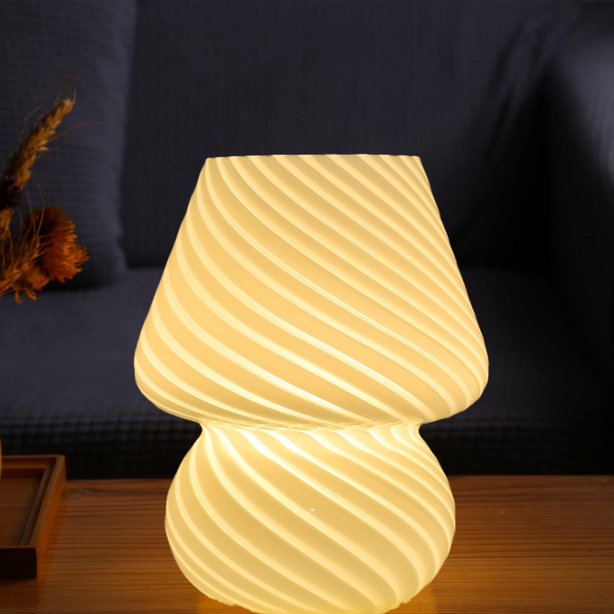 Eine pilzförmige Lampe mit Wirbeldesign. Die Lampe steht auf einem Holztisch. Im Hintergrund ein dunkelblaues Sofa und Trockenblumen.