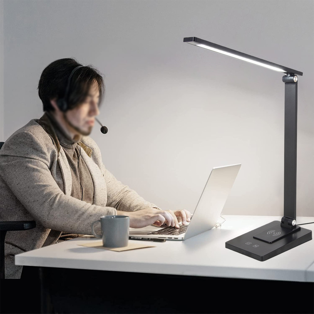 Eine Person mit Headset arbeitet an einem Laptop an einem Schreibtisch, der von einer schwarzen Schreibtischlampe beleuchtet wird.