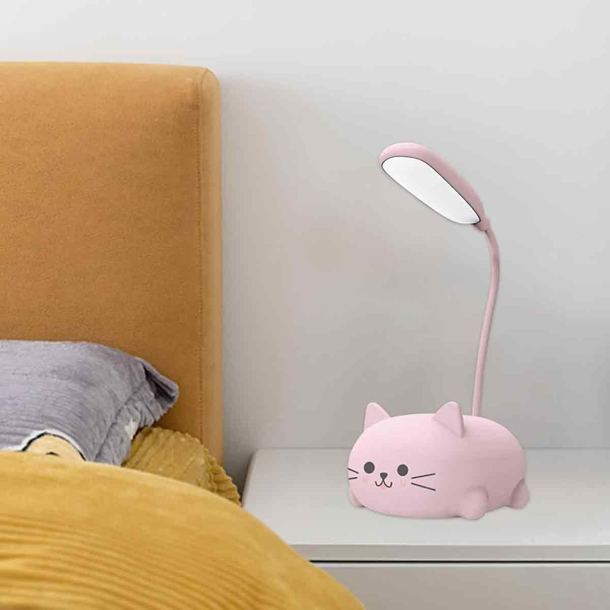 Eine rosa katzenförmige Schreibtischlampe mit flexiblem Hals steht auf einer weißen Oberfläche. Im Hintergrund befindet sich ein Bett mit gelben und grauen Bettwaren.