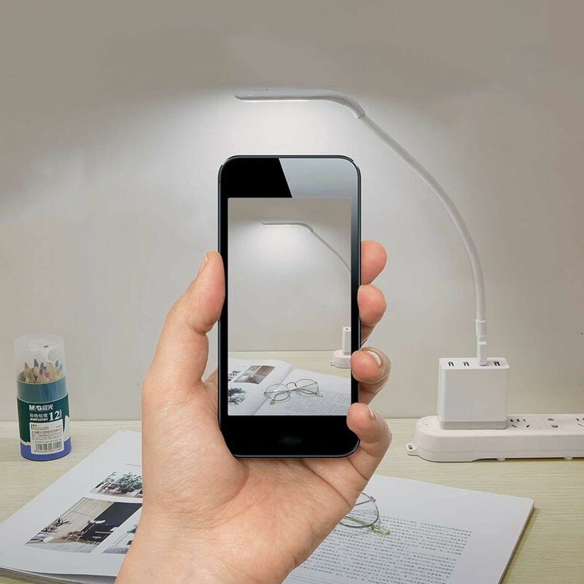 Eine Hand hält ein Telefon, das eine Schreibtischlampe zeigt. Auf dem Tisch befinden sich ein Federmäppchen, ein offenes Buch und eine Brille. Eine Steckdose ist vorhanden.
