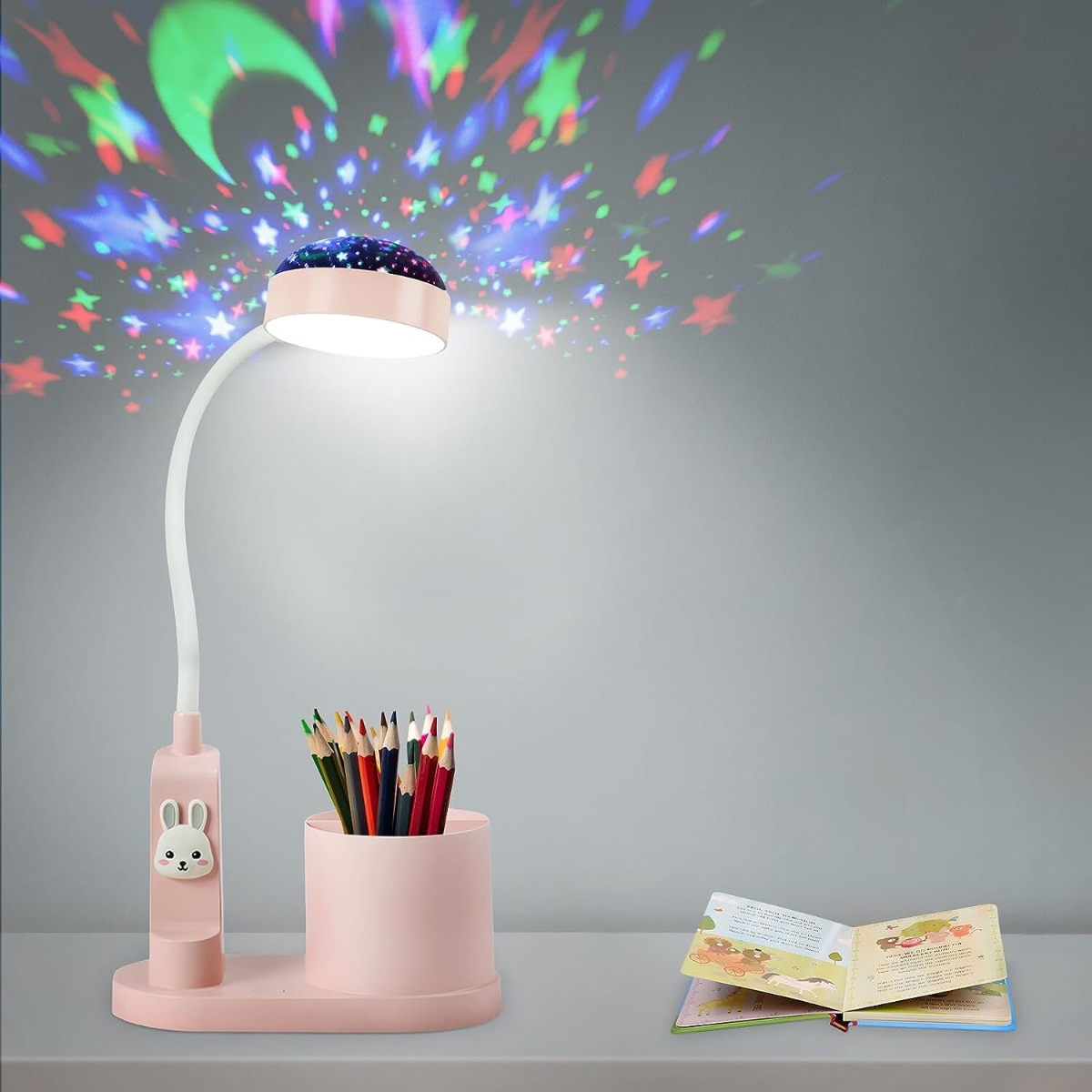 Rosa Schreibtischlampe mit Stern- und Mondprojektor. Stifte und ein Buch liegen auf dem Tisch. Die Wand hat Sternprojektionen.
