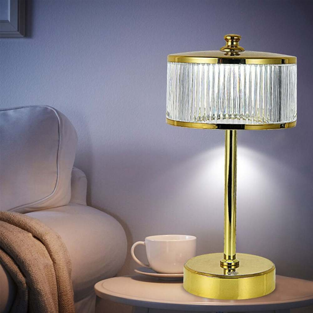 Eine goldene Lampe mit Kristallschirm auf einem Tisch neben einer Teetasse, Couch und Decke. Die Lampe ist beleuchtet.