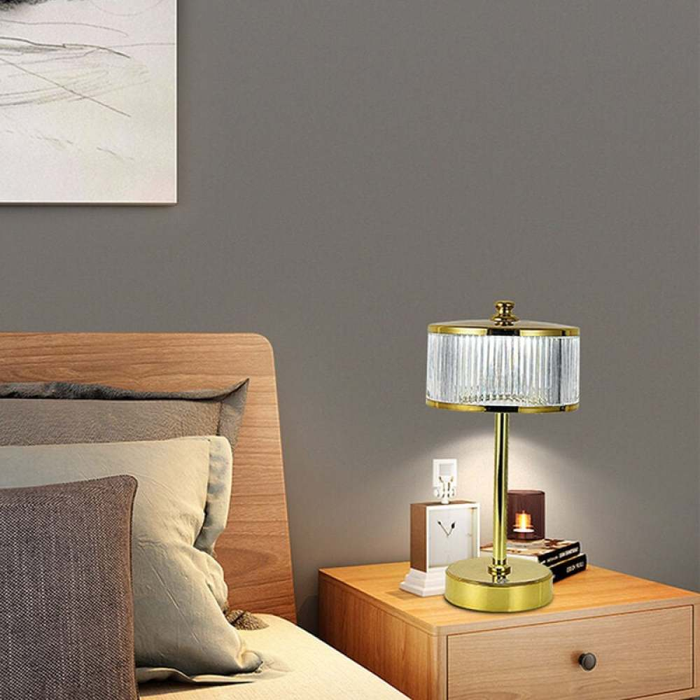 Eine goldene Lampe mit geripptem Glasschirm steht auf einem Nachttisch. Im Hintergrund ist ein Bett, darüber ein Gemälde.