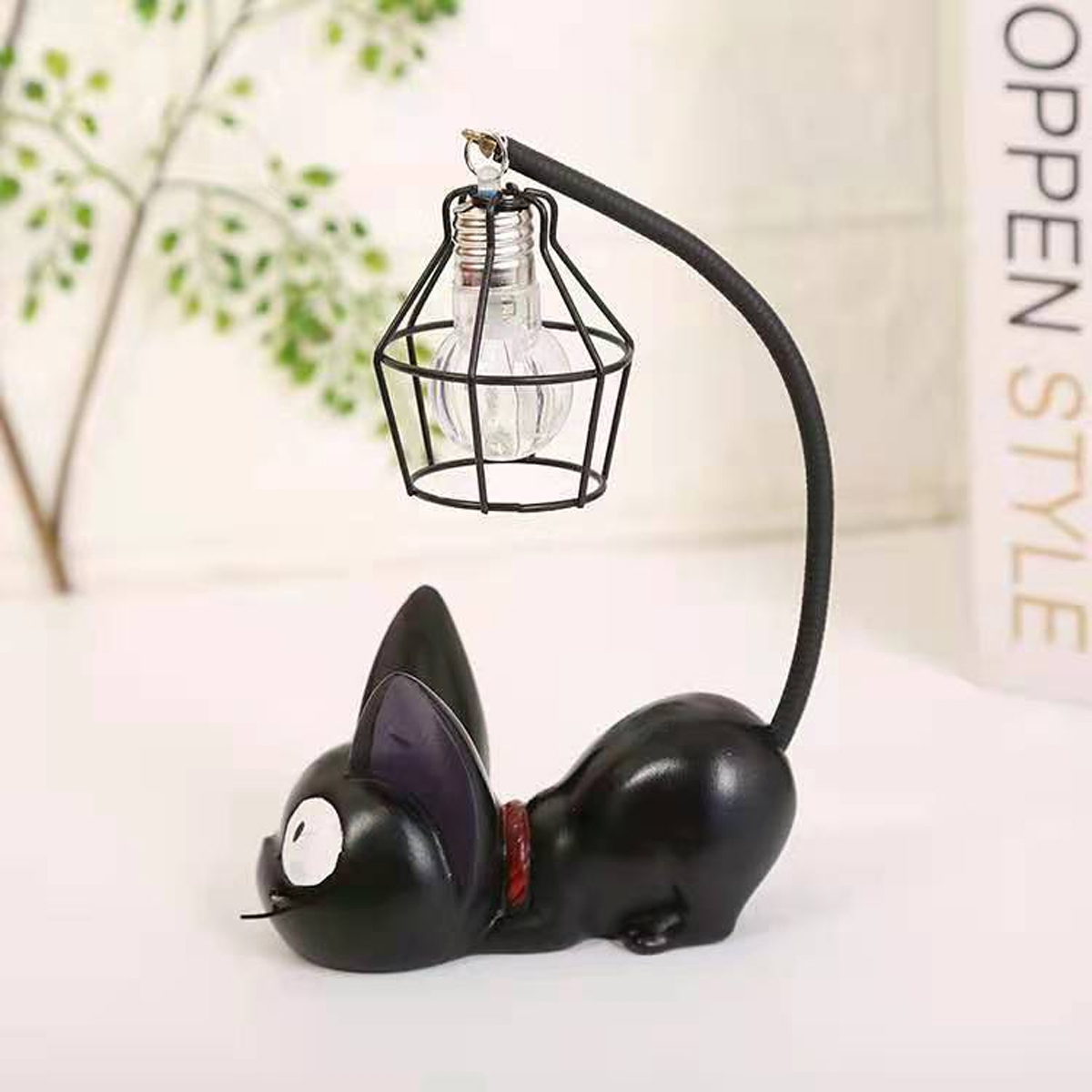 Eine schwarze Katzenlampe mit einem käfigartigen Lampenschirm. Die Katze hat ein rotes Halsband. Der Hintergrund umfasst eine Pflanze und ein Buch.