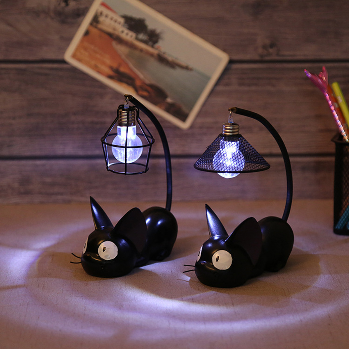 Zwei schwarze Katzenlampen mit leuchtenden Glühbirnen. Im Hintergrund eine Postkarte und Stifte. Die Lampen stehen auf einem Tisch.