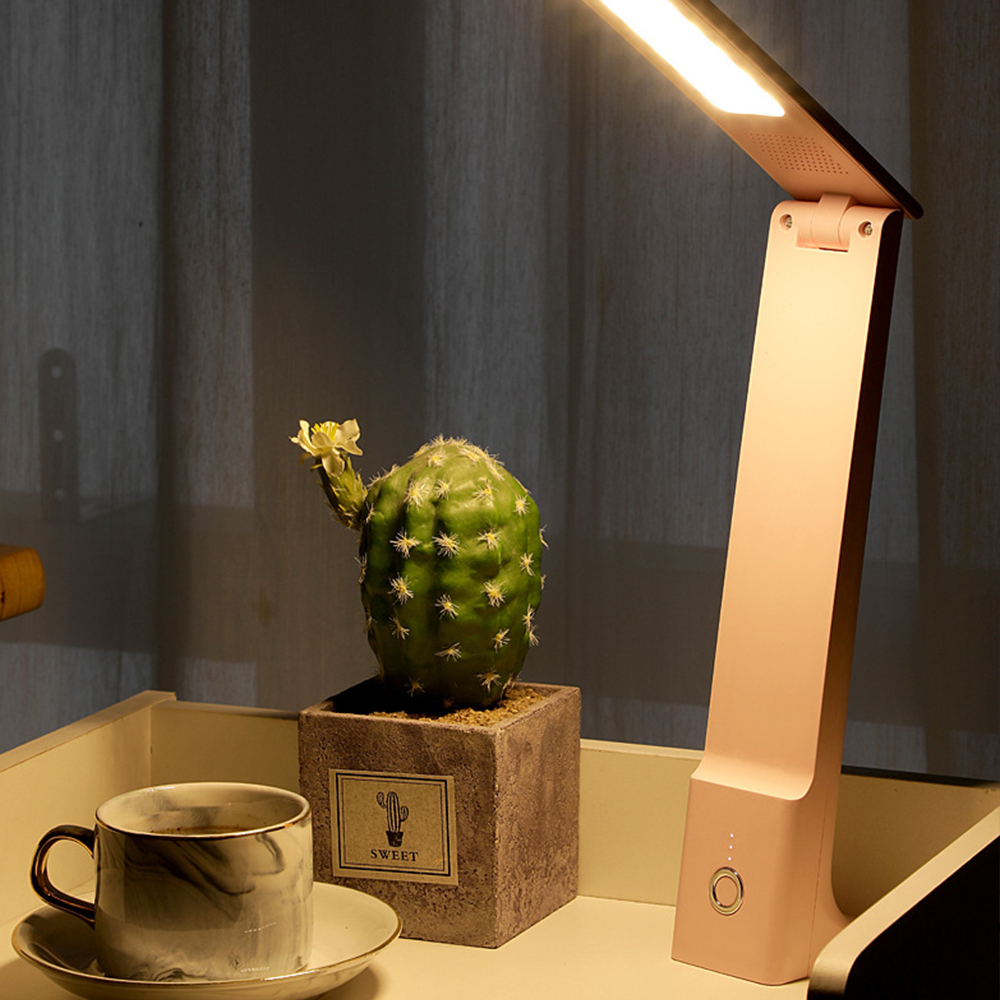 Ein Schreibtisch mit einer rosa Lampe, einem Kaktus und einer Kaffeetasse. Die Lampe ist an. Im Hintergrund ist ein Vorhang.