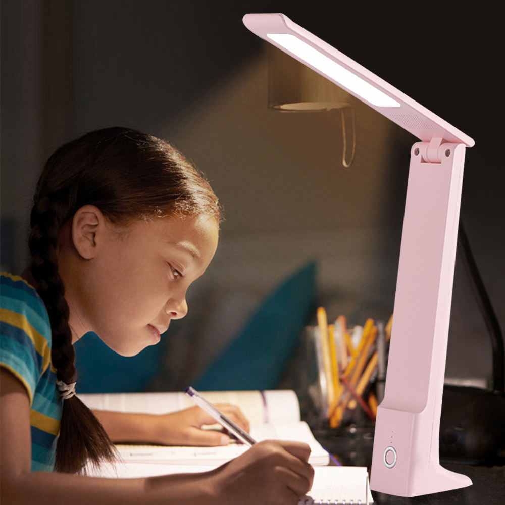 Ein Mädchen schreibt in einem Notizbuch unter einer rosa Schreibtischlampe. Die Lampe ist an und beleuchtet den Arbeitsbereich.