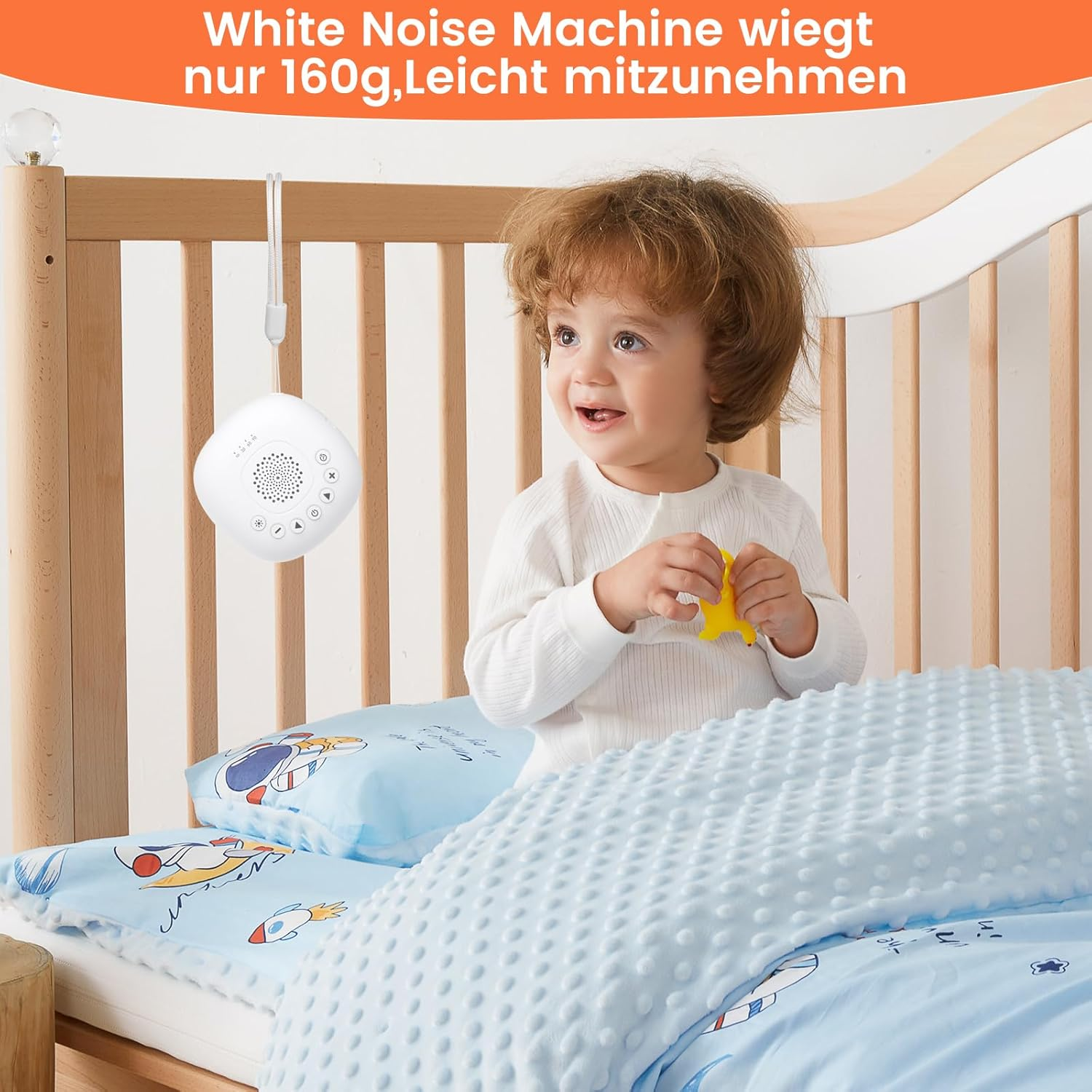 Ein Baby in einem Kinderbett hält ein gelbes Spielzeug. Eine Weißgeräuschmaschine hängt darüber. Blaue Bettwäsche mit Raketendesign ist sichtbar.