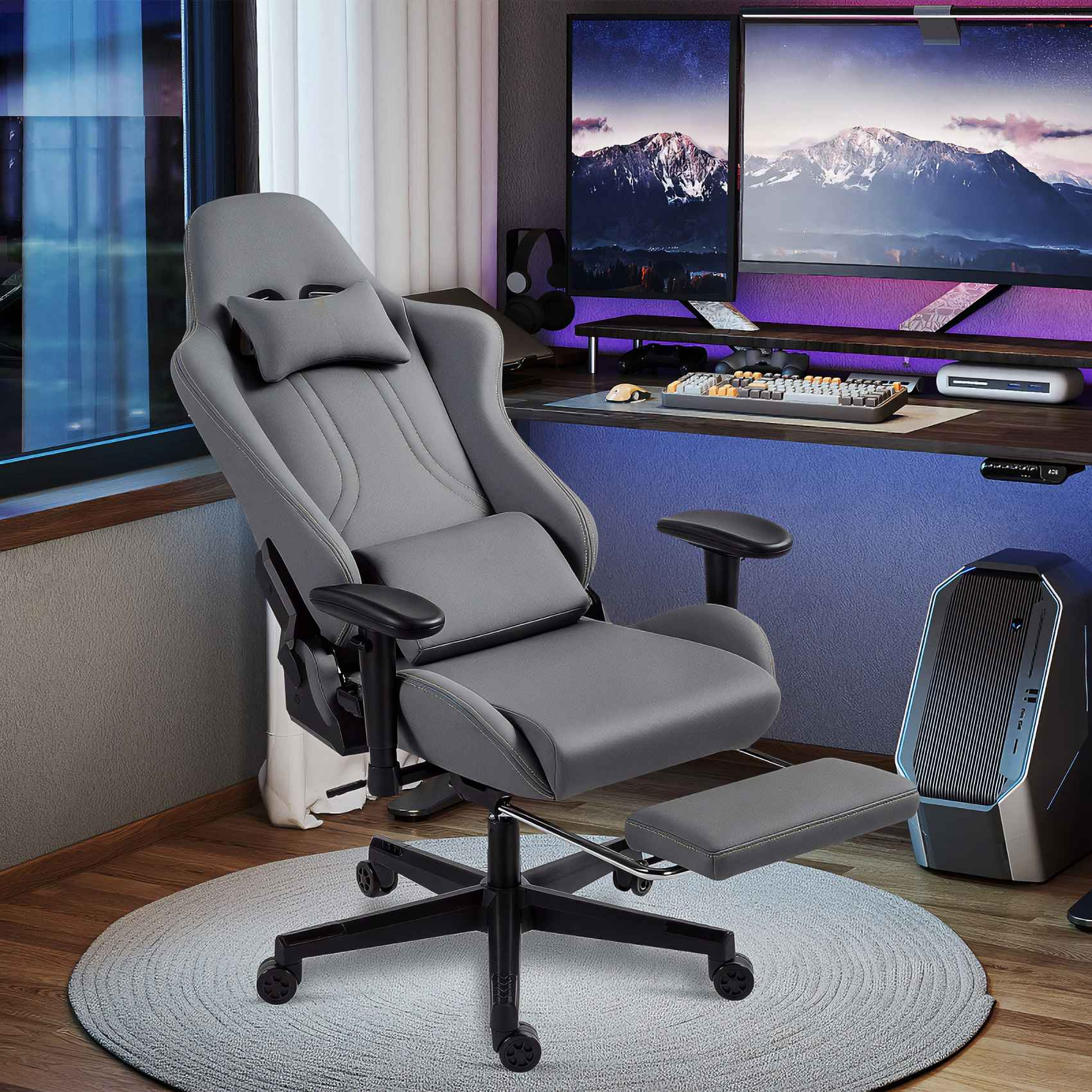 Ein grauer Gaming-Stuhl mit Fußstütze steht in einem Raum mit Schreibtisch, Monitoren und Computer. Im Hintergrund ist ein Fenster.