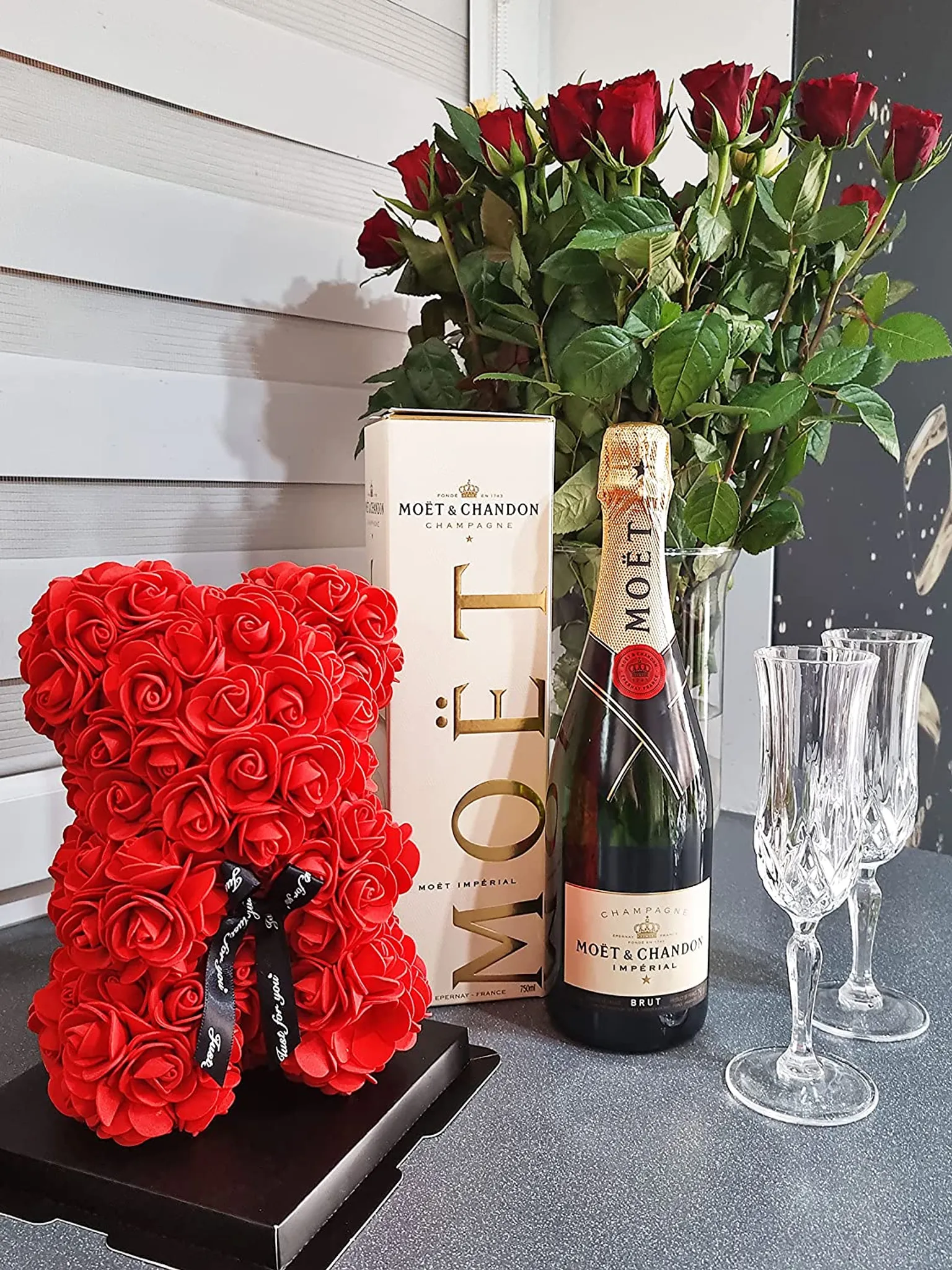 Roter Rosenbär, Champagnerflasche, Rosen in einer Vase und zwei Champagnergläser auf einem Tisch.