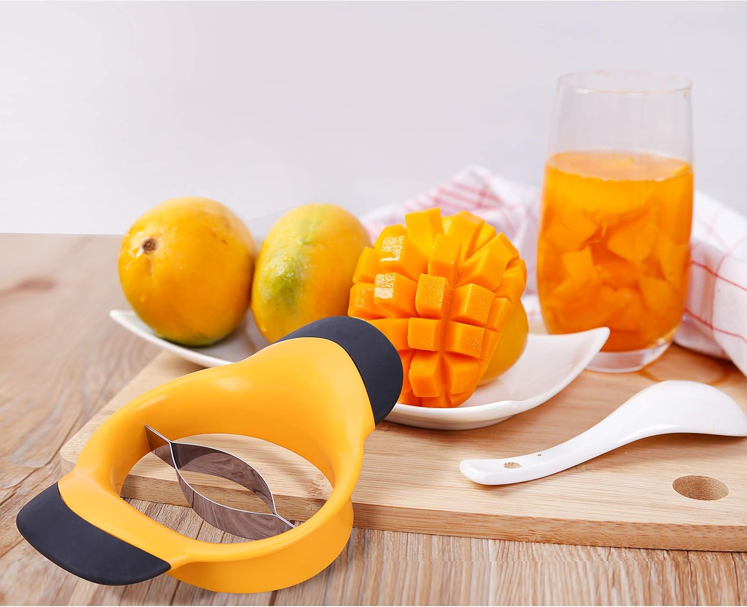 Ein Mangoschneider, Mangos und ein Glas Mangosaft auf einem Holzbrett. Ein Löffel und ein Handtuch sind ebenfalls vorhanden.