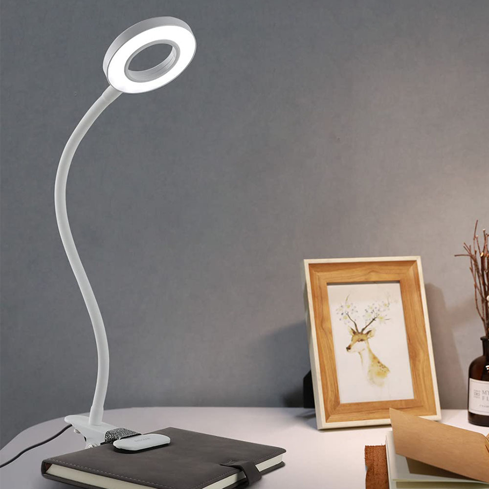 Eine weiße Schreibtischlampe mit kreisförmigem Licht. Ein gerahmtes Hirschbild, ein Notizbuch und eine Vase stehen auf dem Tisch. Graue Wand im Hintergrund.