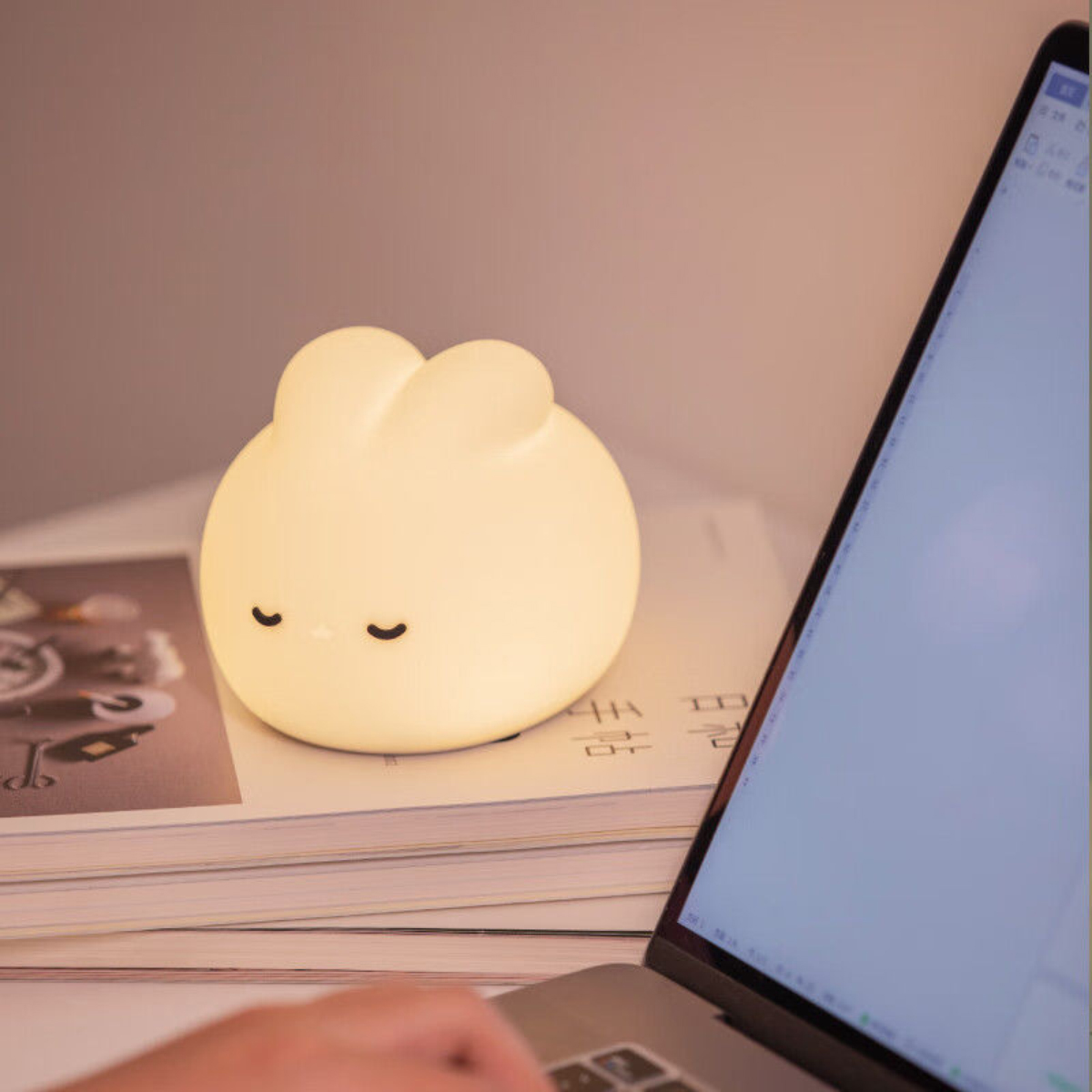 Eine weiche, leuchtende Hasenlampe steht auf Büchern neben einem geöffneten Laptop. Die Lampe hat geschlossene Augen, eine Hand ist sichtbar.