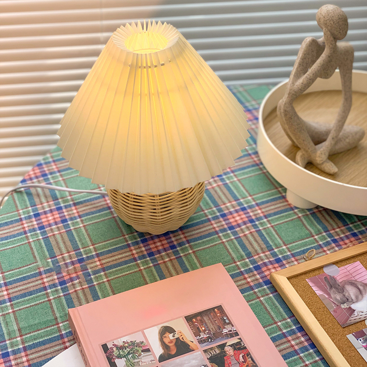 Eine beleuchtete Lampe auf einer karierten Oberfläche mit einem rosa Buch und einer Figur auf einem Tablett. Eine Pinnwand mit einem Bild ist ebenfalls sichtbar.