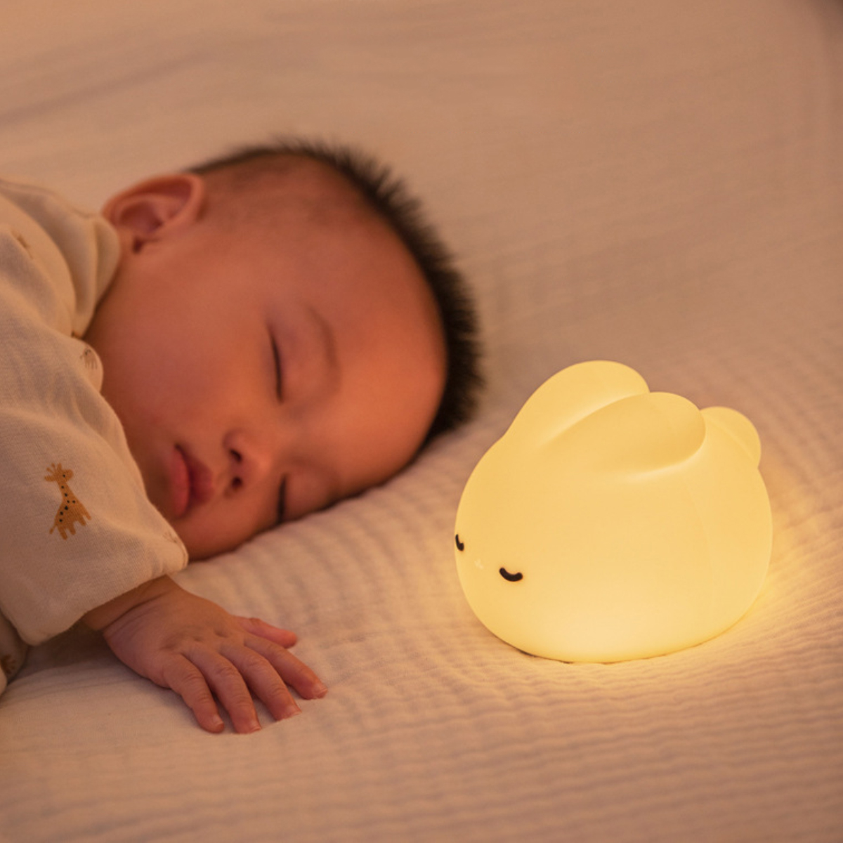 Ein Baby schläft auf einem weißen Laken neben einem leuchtenden, hasenförmigen Nachtlicht. Das Baby trägt einen hellen Strampler.