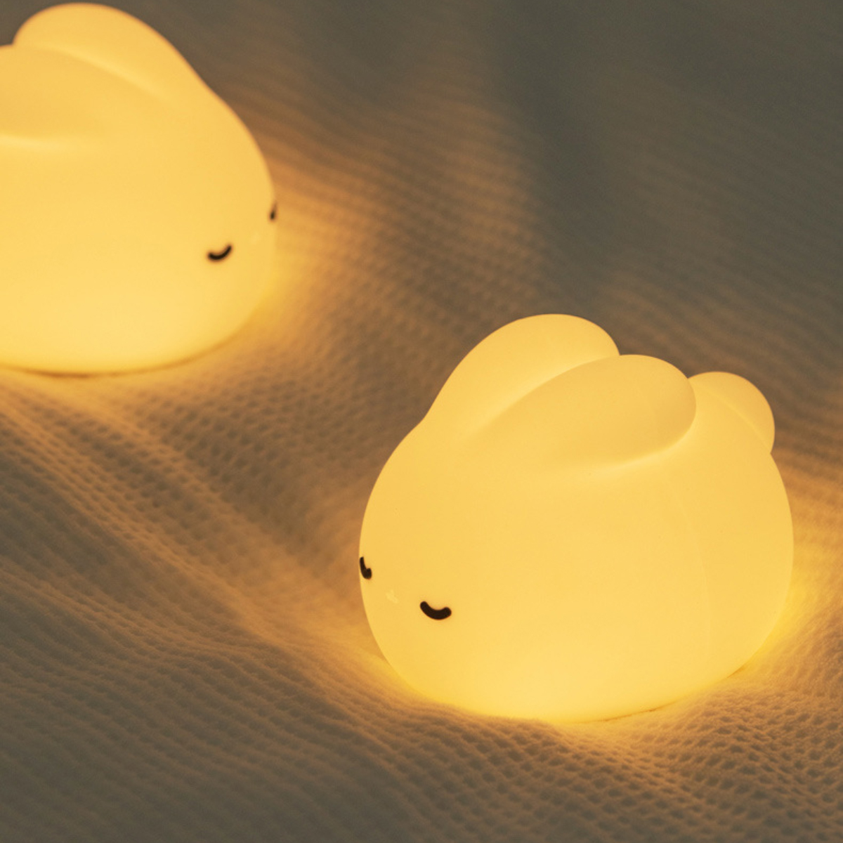 Zwei leuchtende Hasenlampen auf einer strukturierten weißen Oberfläche. Jede Lampe hat geschlossene Augen und strahlt ein sanftes gelbes Licht aus.