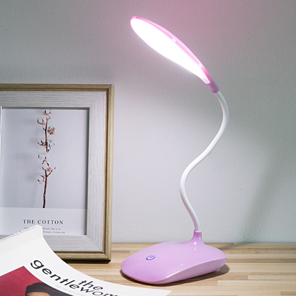 Eine rosa Schreibtischlampe steht auf einer Holzoberfläche. Ein gerahmtes Bild und eine Zeitschrift sind in der Nähe. Die Lampe leuchtet.